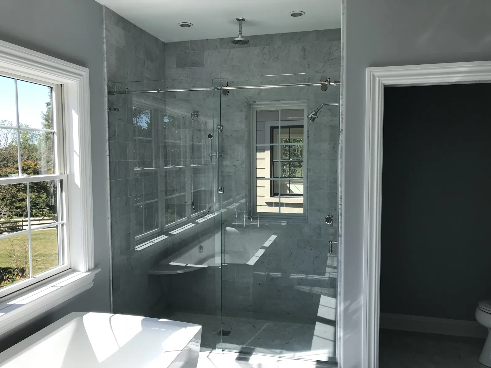 Modern bathroom with glass-enclosed shower, marble tiles, bathtub, and large windows with natural light.