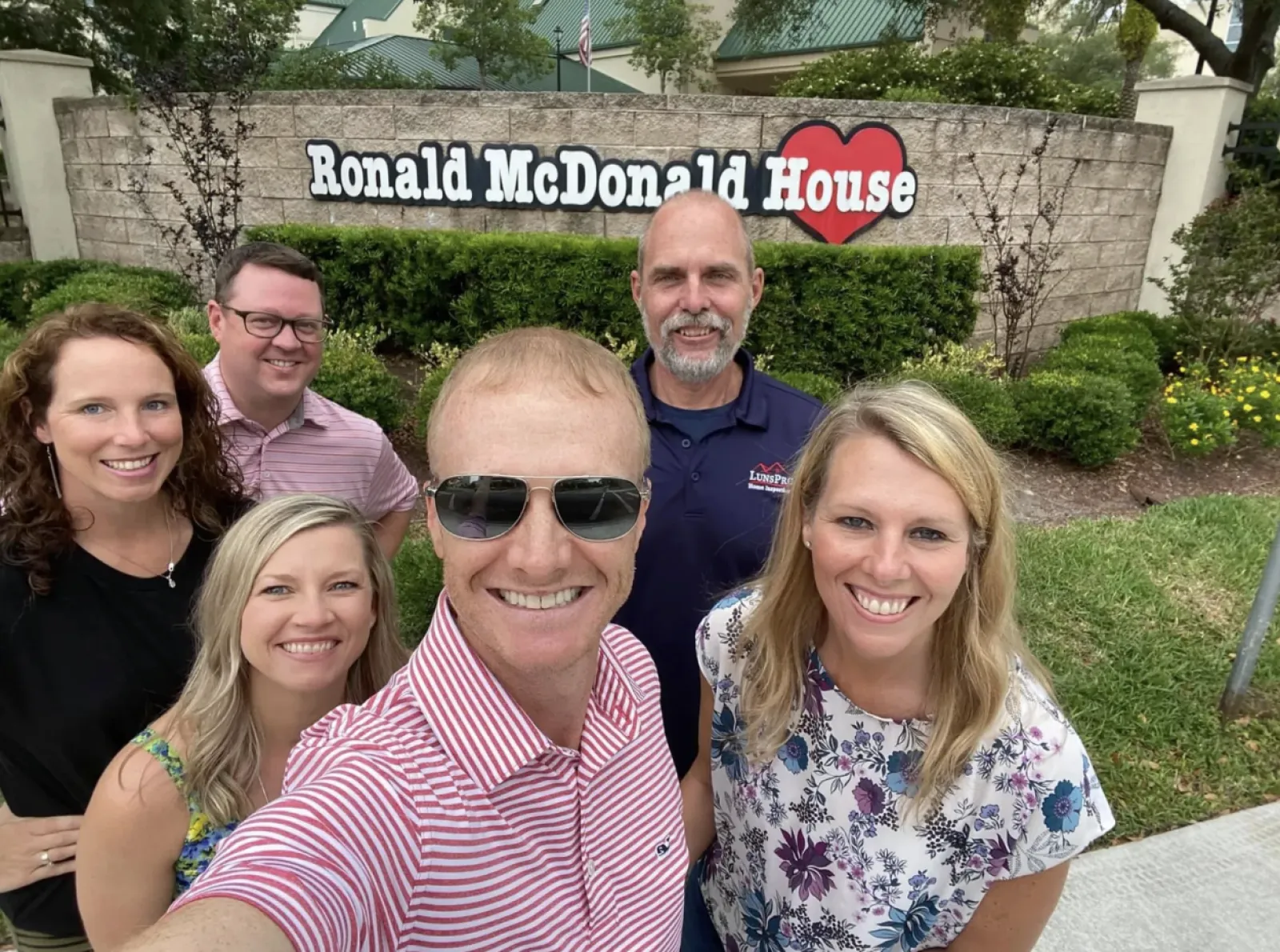 Group selfie of six smiling adults in front of Ronald McDonald House sign outdoors on a sunny day
