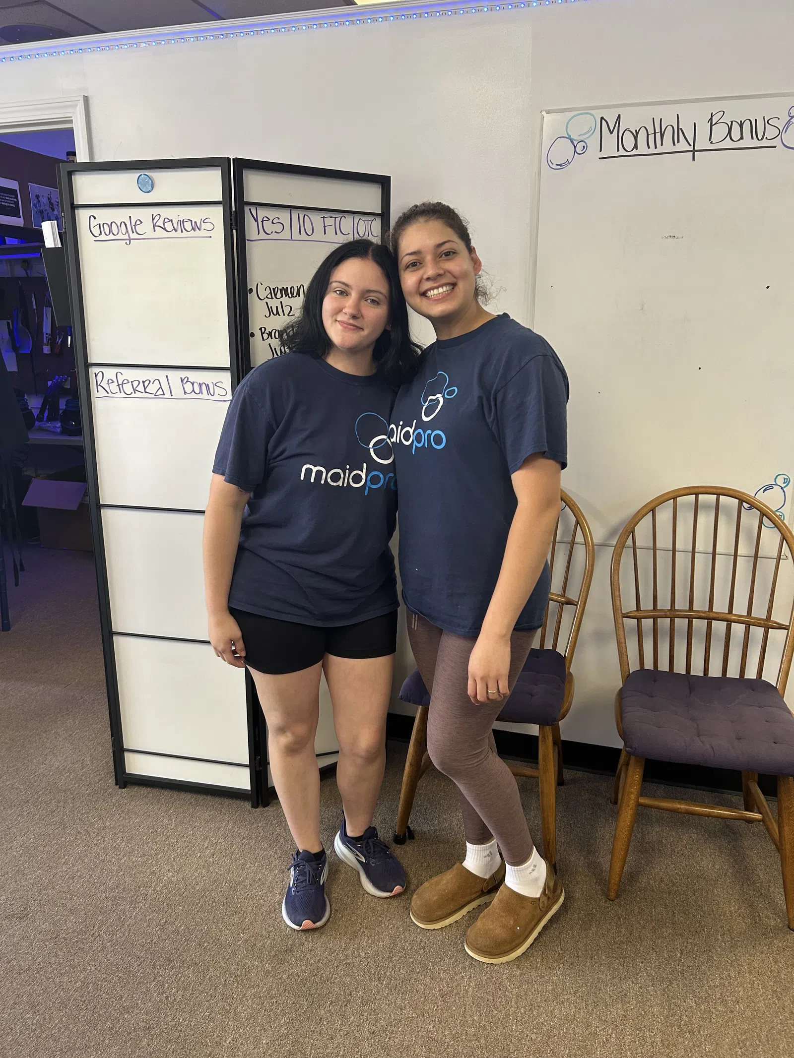 Two women smiling wearing MaidPro t-shirts standing in front of office whiteboards and chairs.