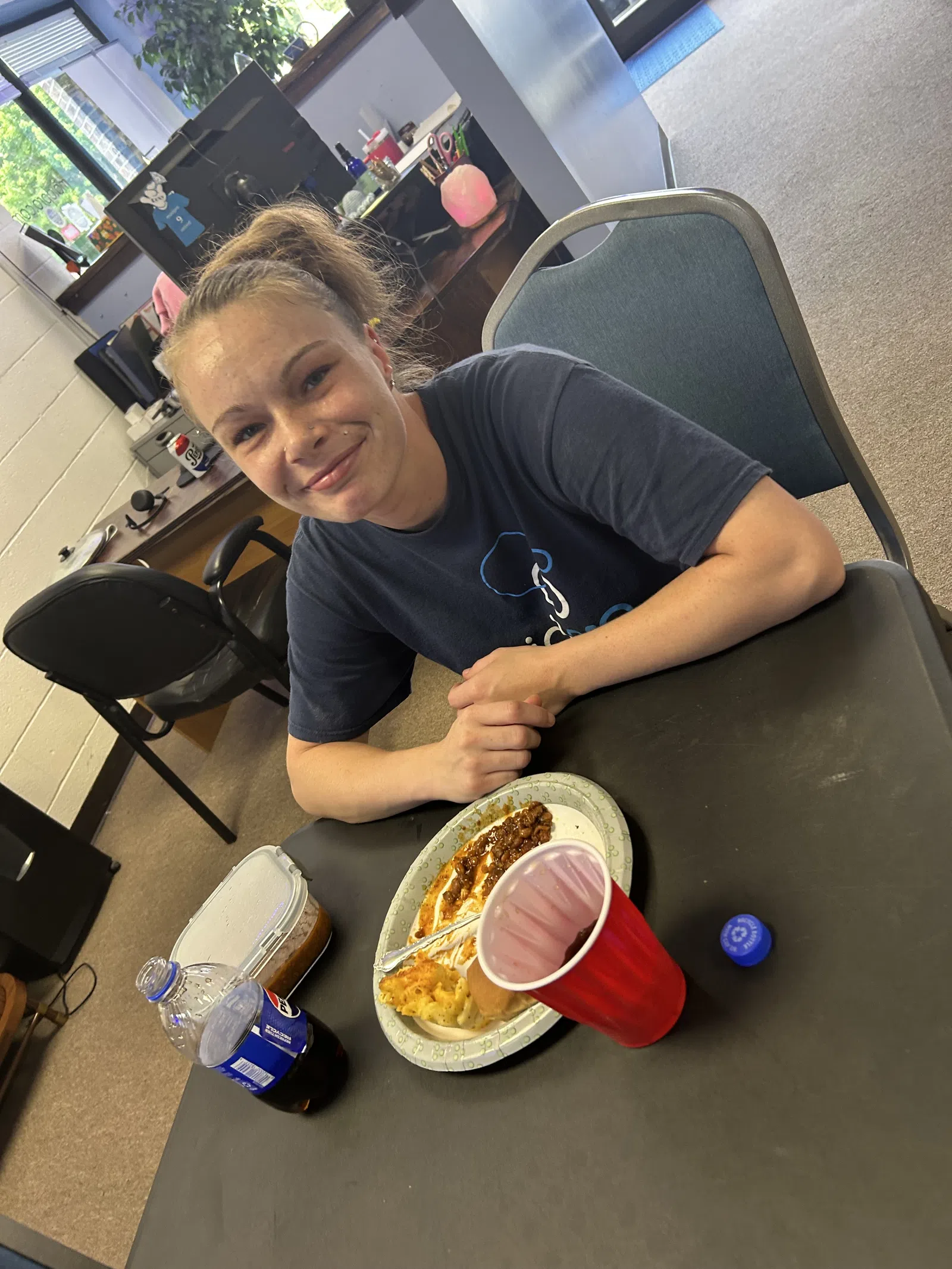 Young woman smiling at a table with a plate of food, a red cup, and a soda bottle in an office setting.