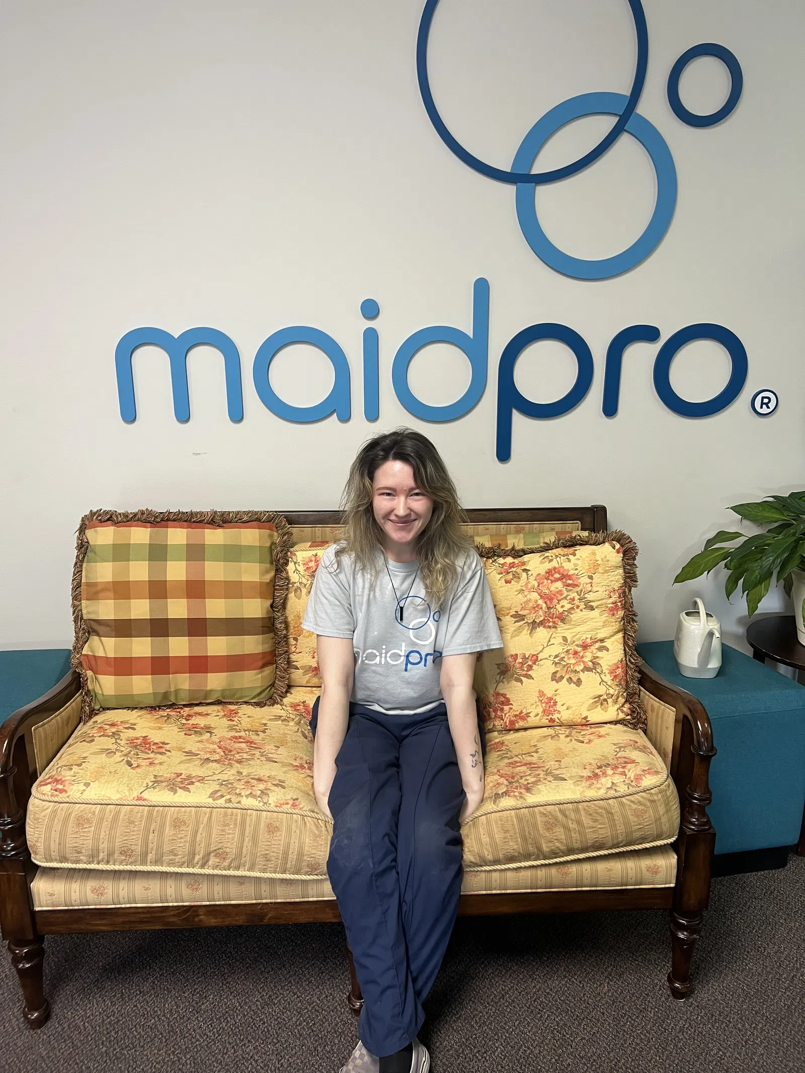 Smiling woman in MaidPro shirt sitting on floral couch under MaidPro logo with decorative pillows and plant nearby
