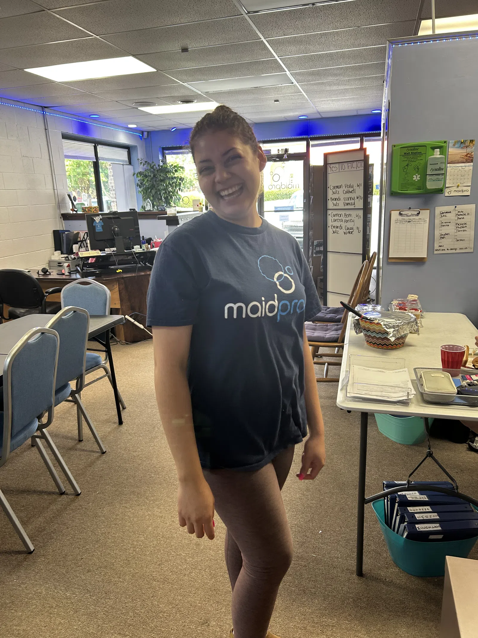 Smiling woman wearing a MaidPro shirt stands in a bright office with chairs, a table, and documents.