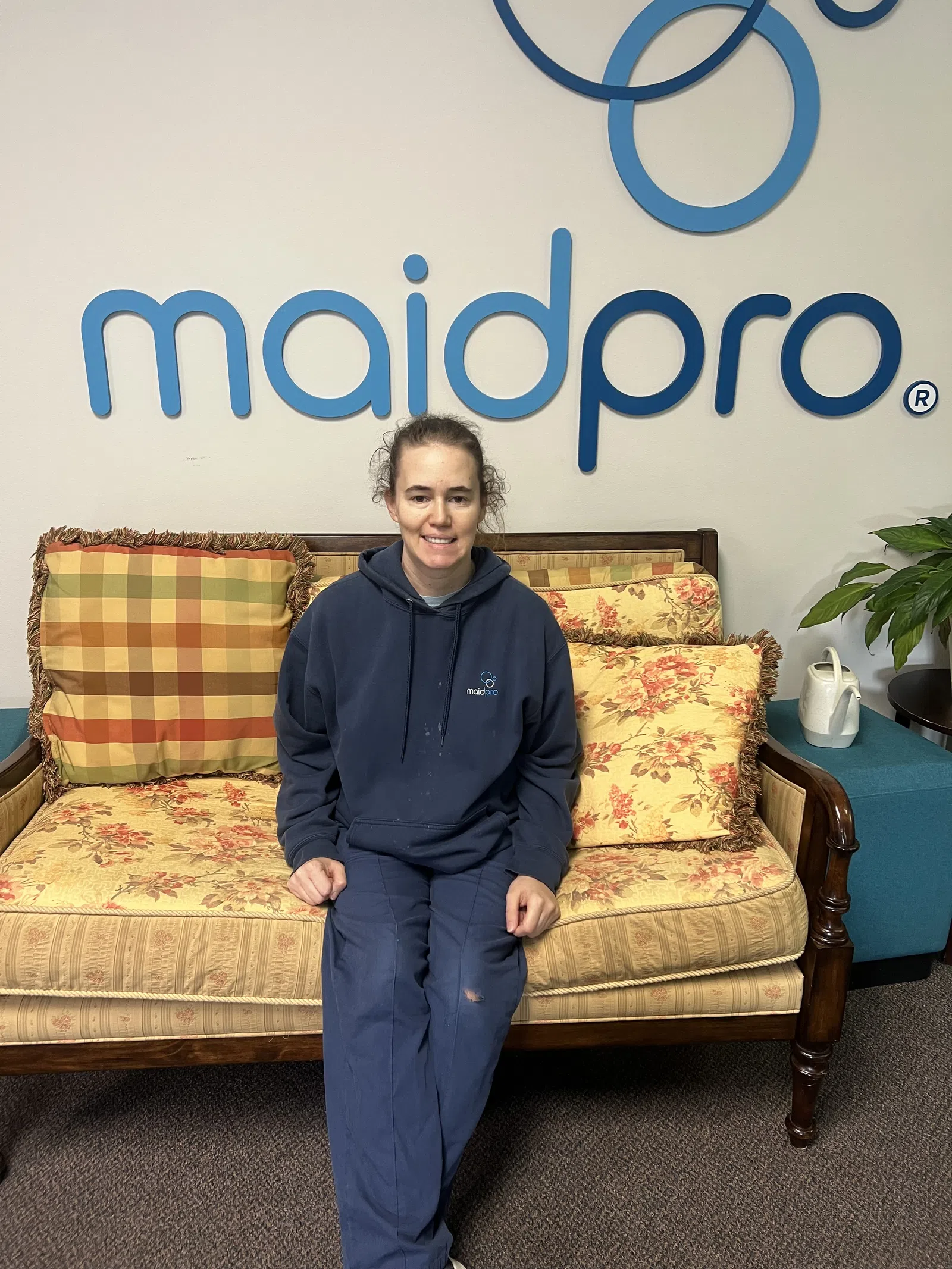 Woman in MaidPro uniform sitting on vintage floral couch in office with MaidPro logo behind her.