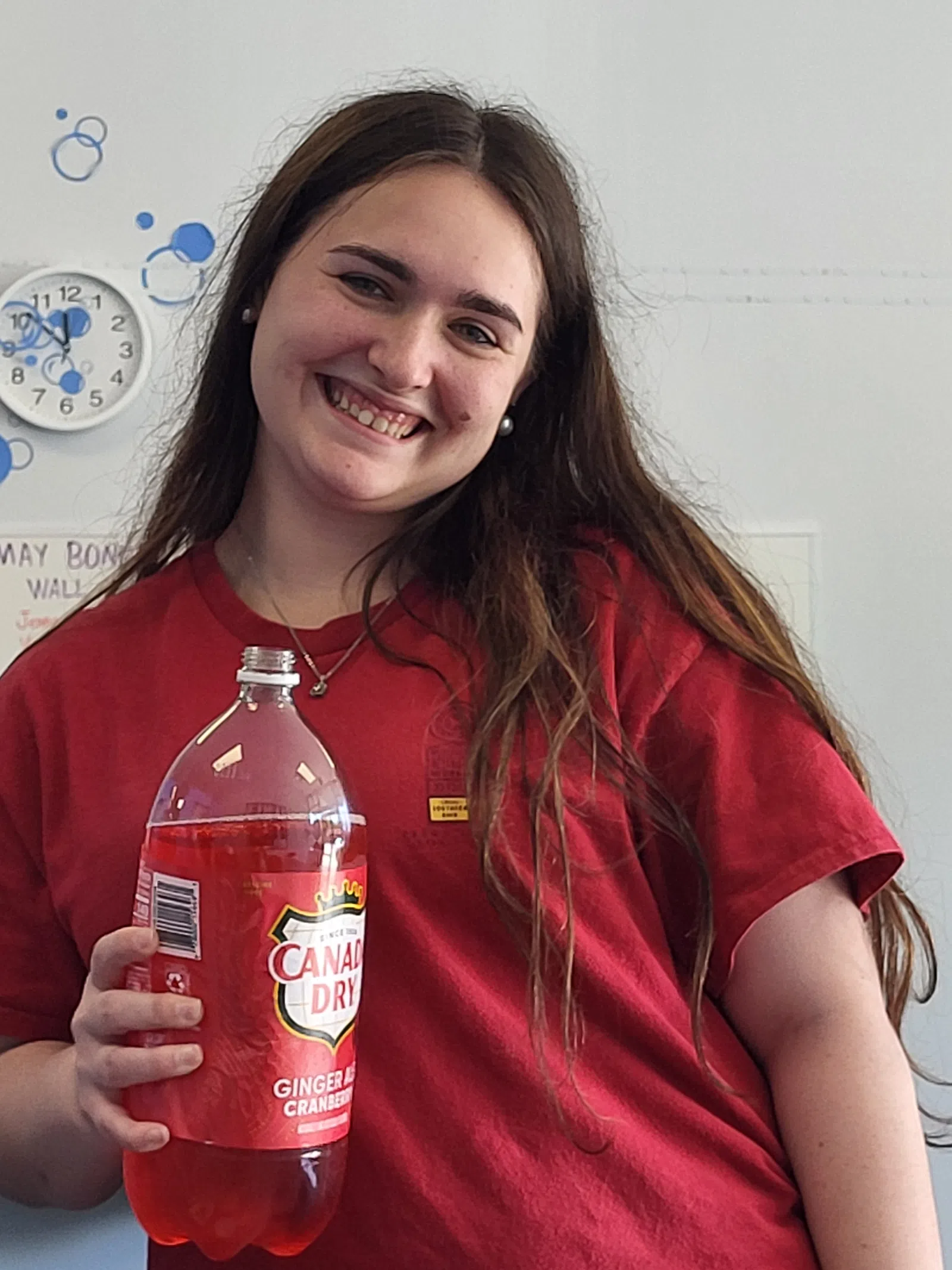 Smiling young woman in red shirt holding a Canada Dry Ginger Ale Cranberry soda bottle indoors.