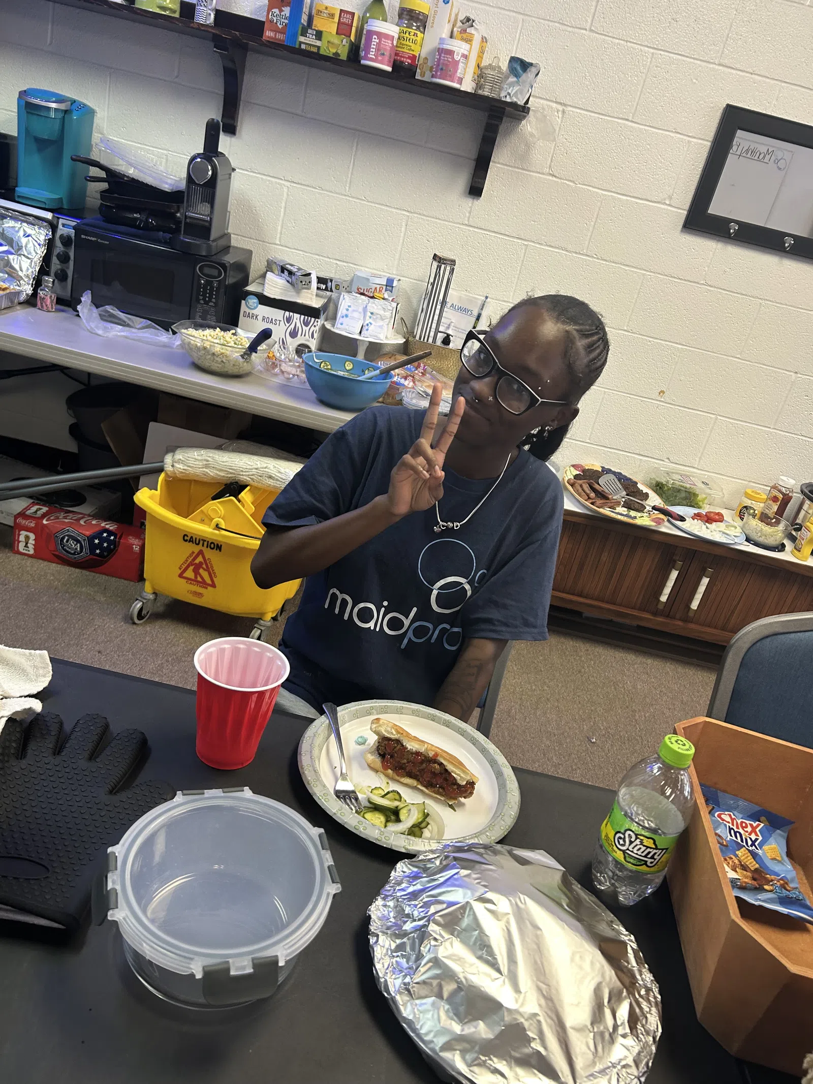 Person wearing MaidPro shirt making peace sign while sitting at table with food and drink in break room.