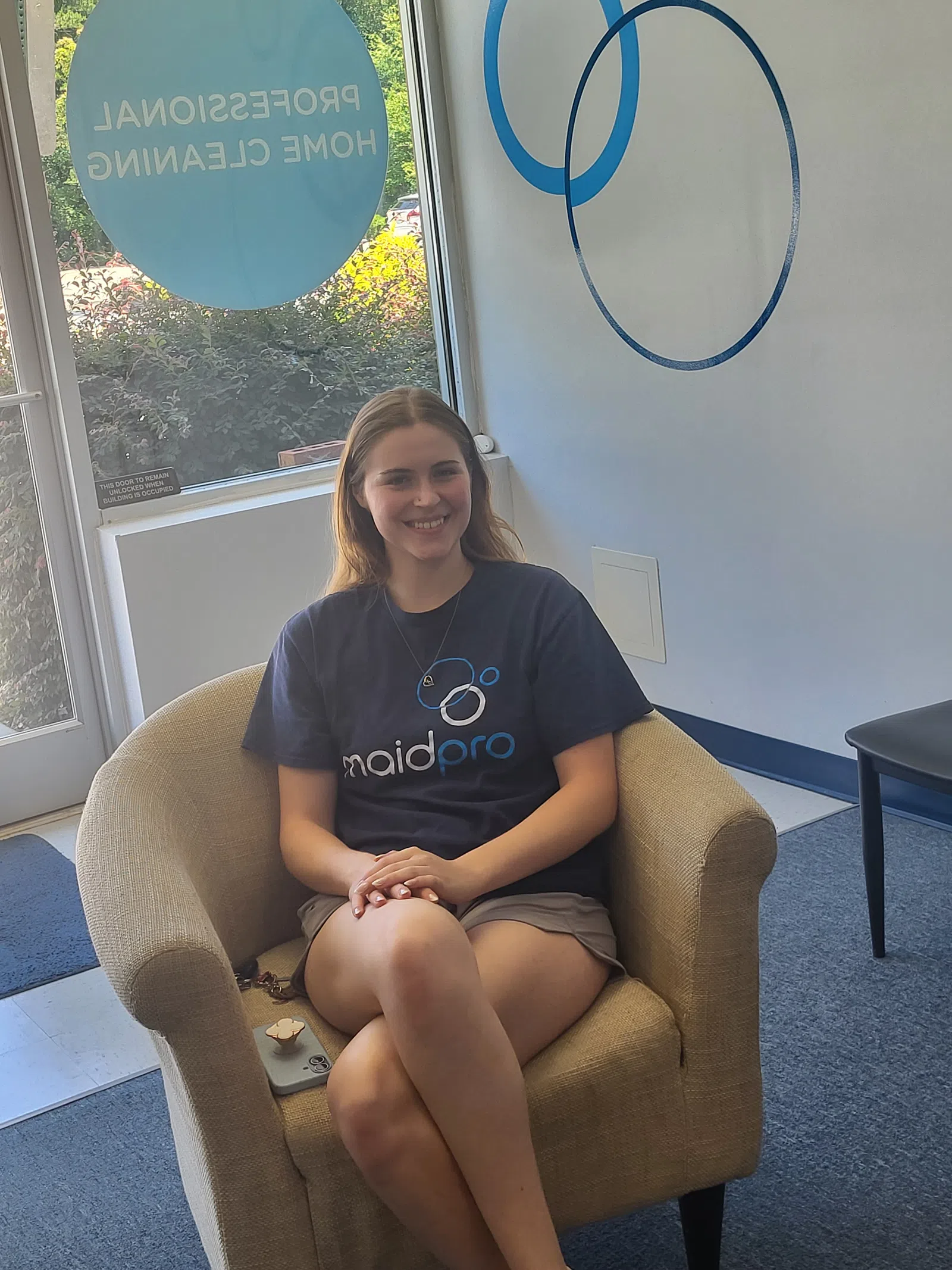 Smiling woman wearing MaidPro shirt sitting comfortably in an armchair in a professional home cleaning office.