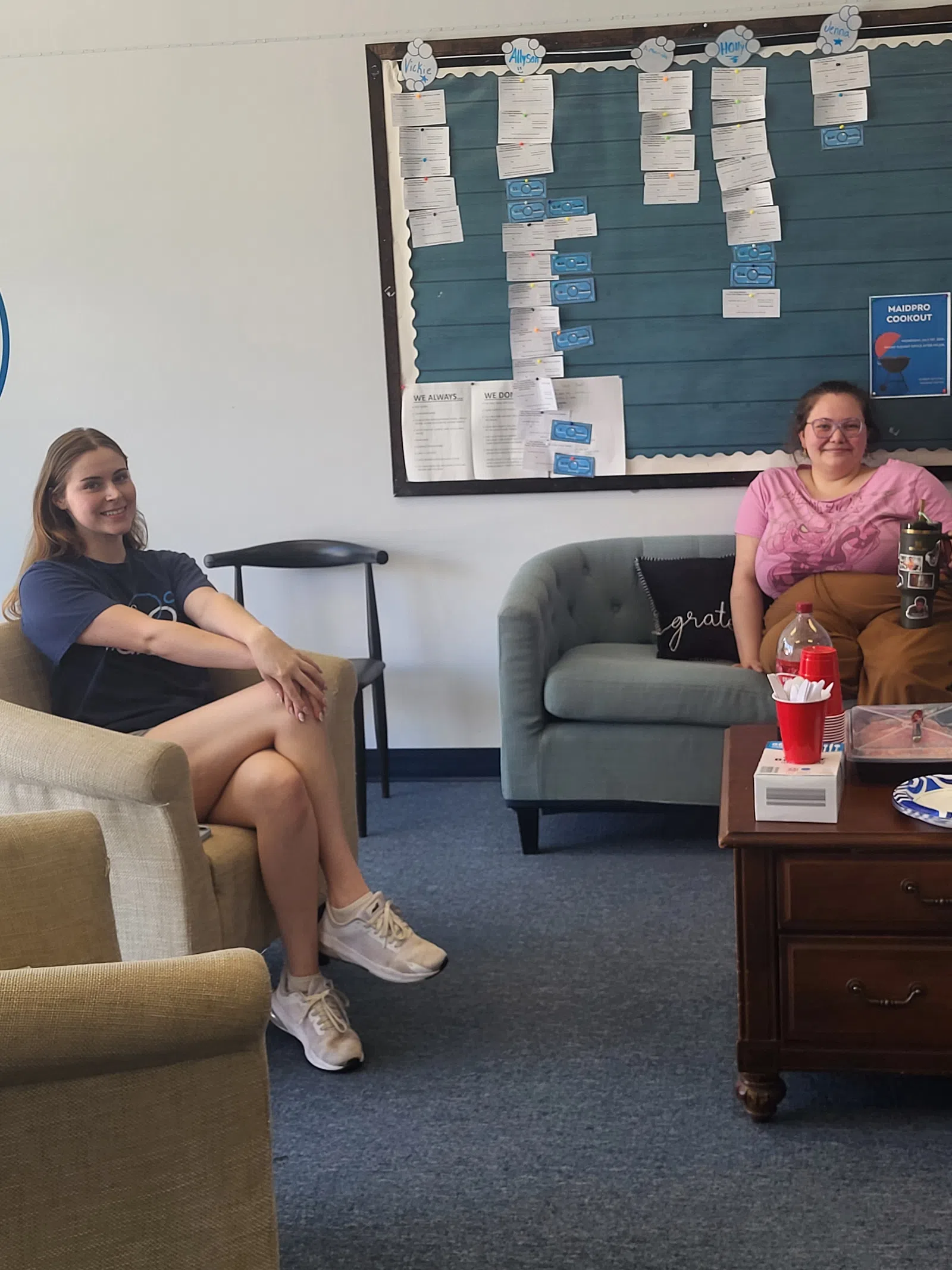 Two women sitting in a casual office lounge with a bulletin board and coffee table with snacks.