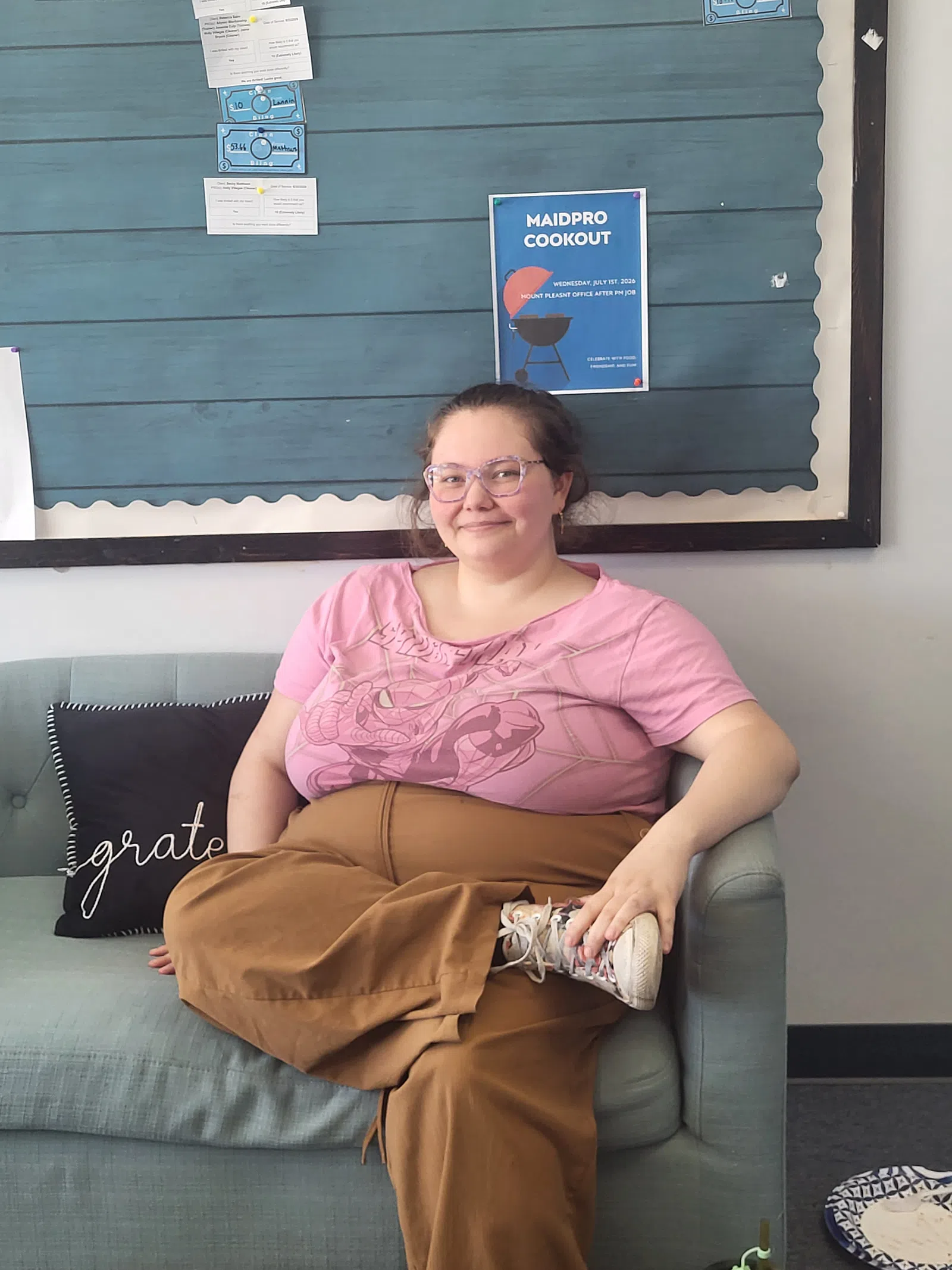 Smiling woman in pink shirt and brown pants sitting cross-legged on light gray couch with bulletin board background.