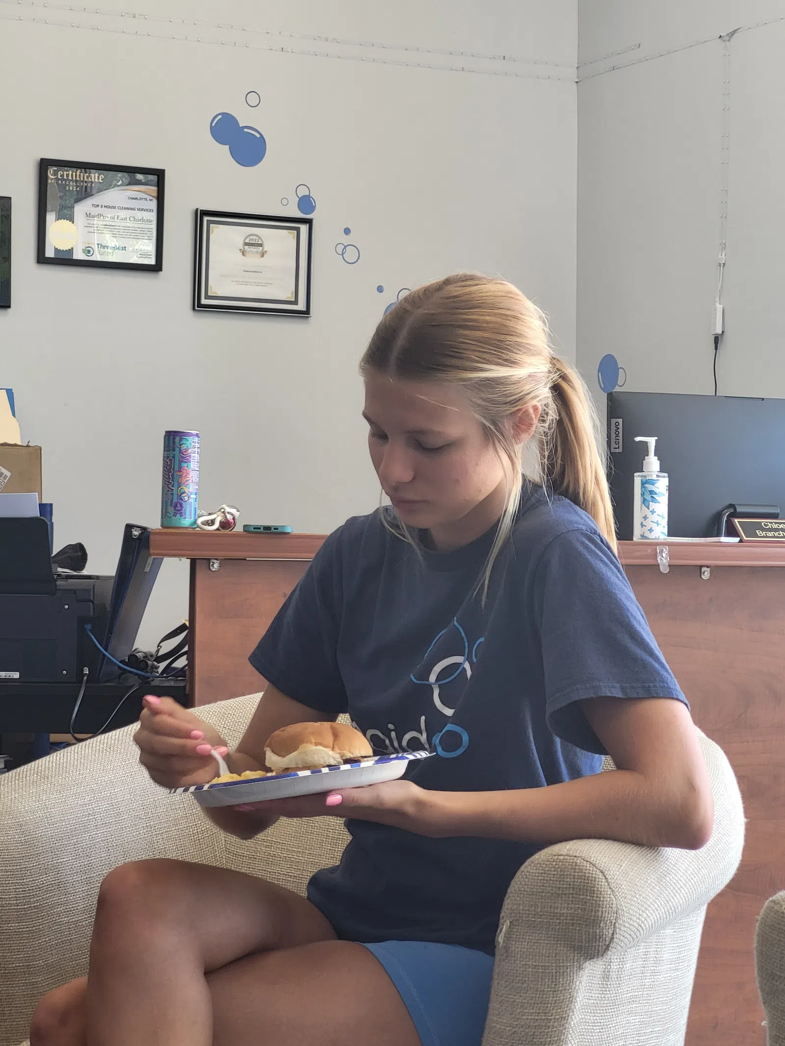 Young woman in navy shirt sitting on beige chair eating from a plate with sandwich and food indoors.