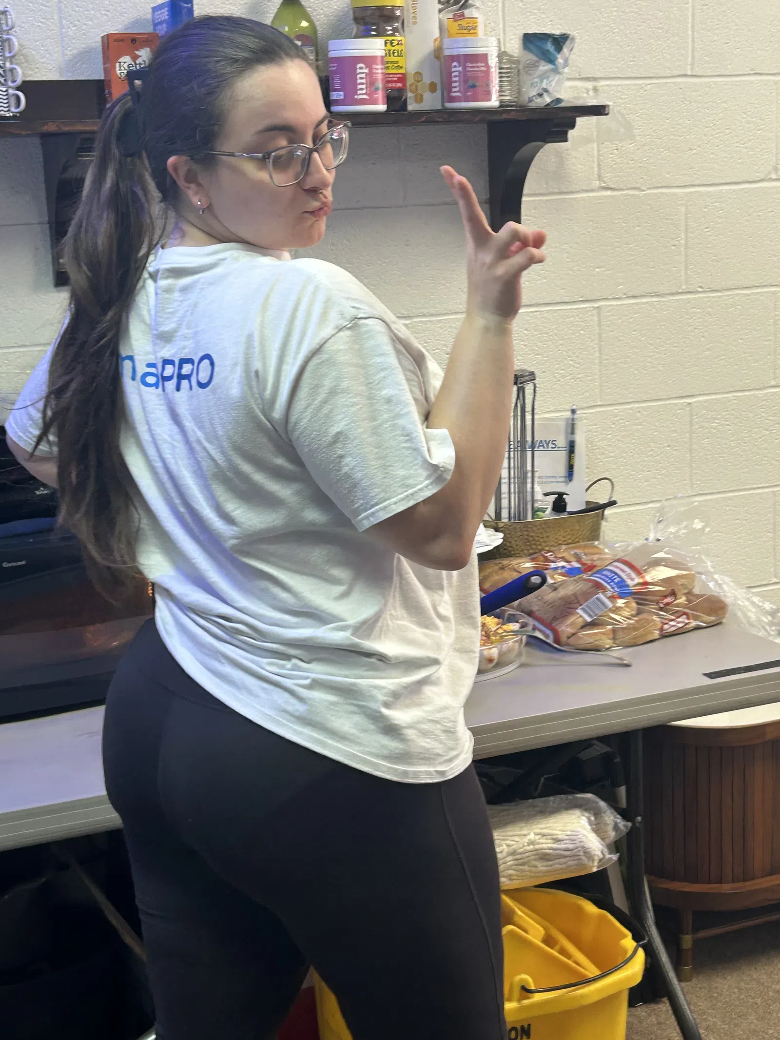 Woman in glasses and white T-shirt posing in a kitchen area with food items and cleaning supplies visible.