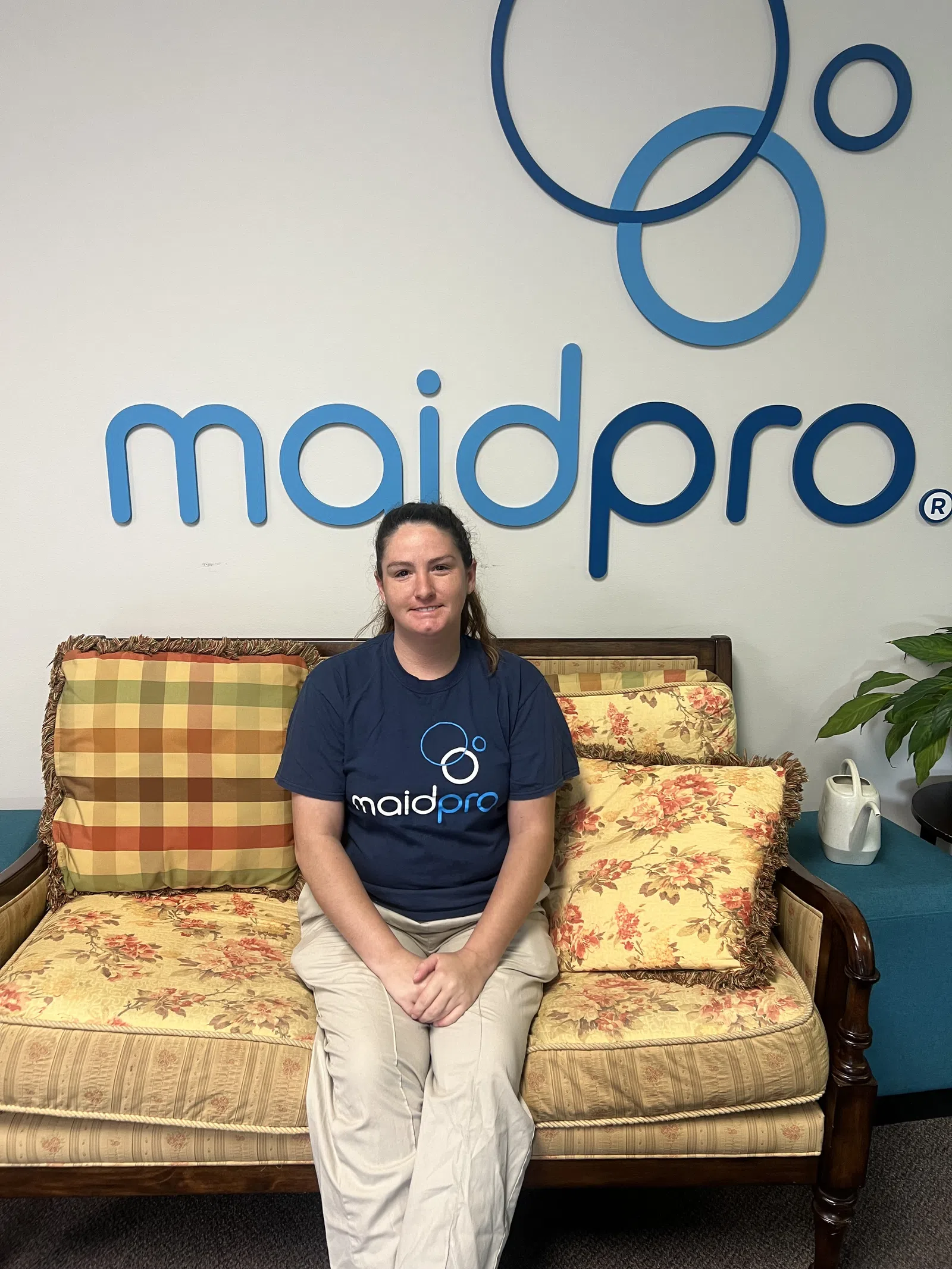 Woman in Maid Pro uniform sitting on floral patterned couch with Maid Pro logo on wall behind her.