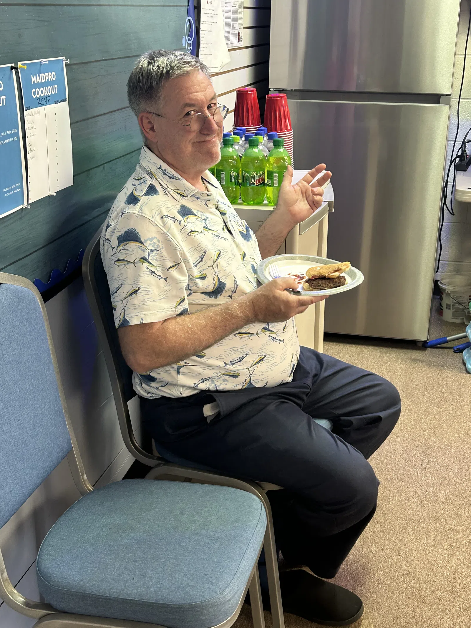 Man with glasses in patterned shirt sitting and eating a burger at an indoor gathering with drinks and red cups nearby.