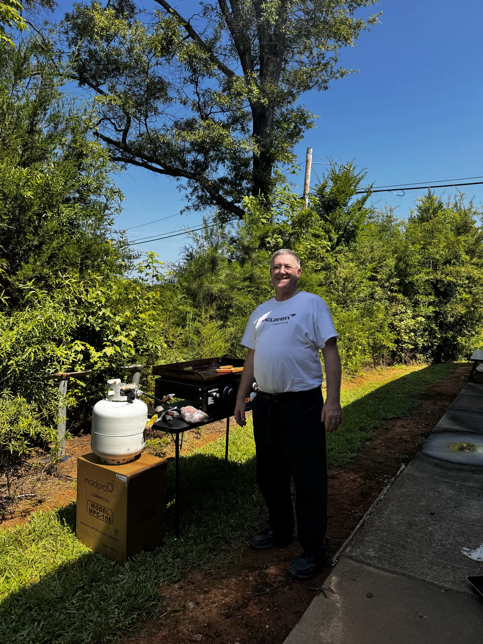 Man standing near a grill with propane tank in a sunny backyard surrounded by green trees and blue sky.