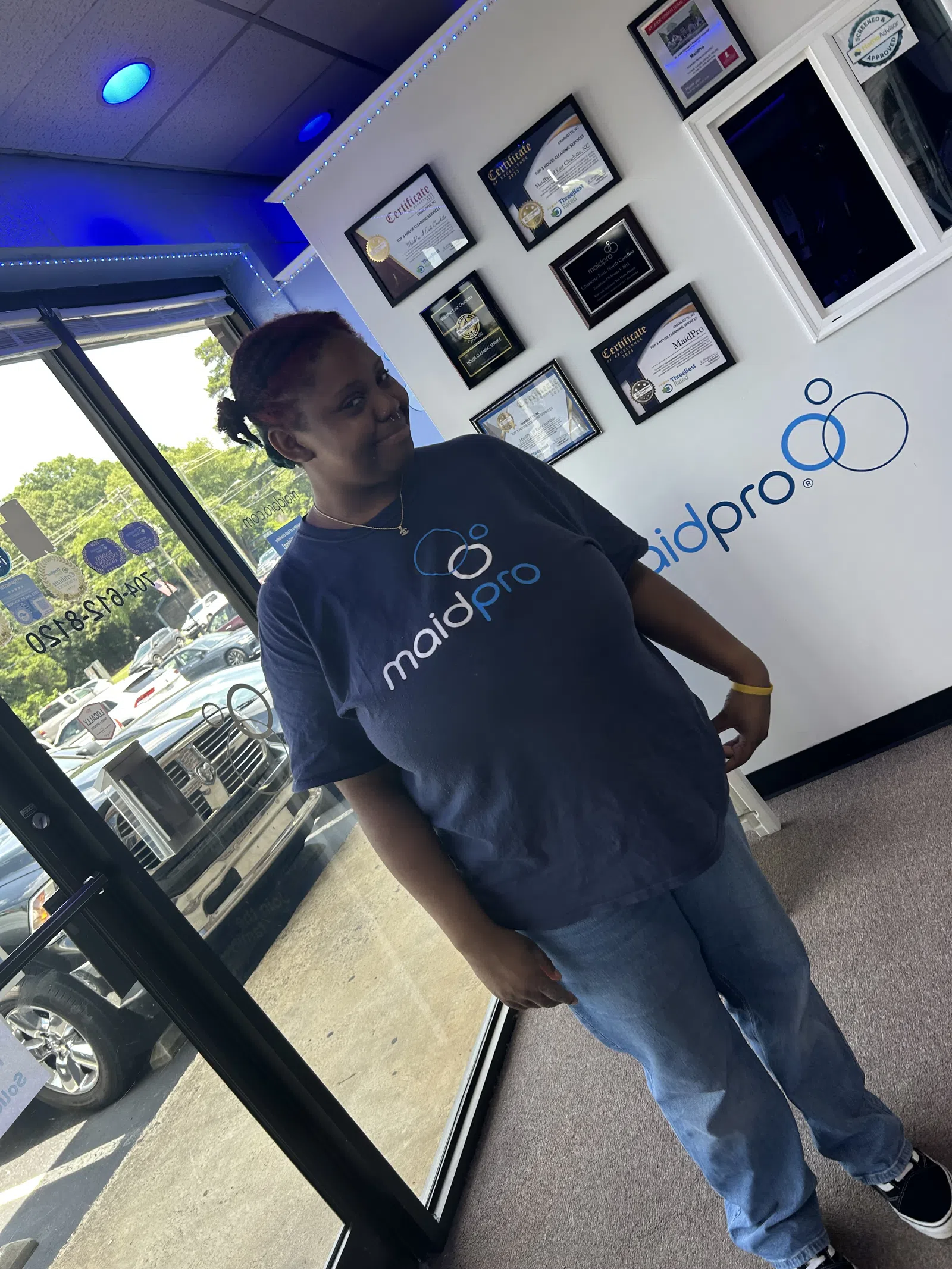 Woman standing inside MaidPro office wearing a MaidPro t-shirt near certificates and window.