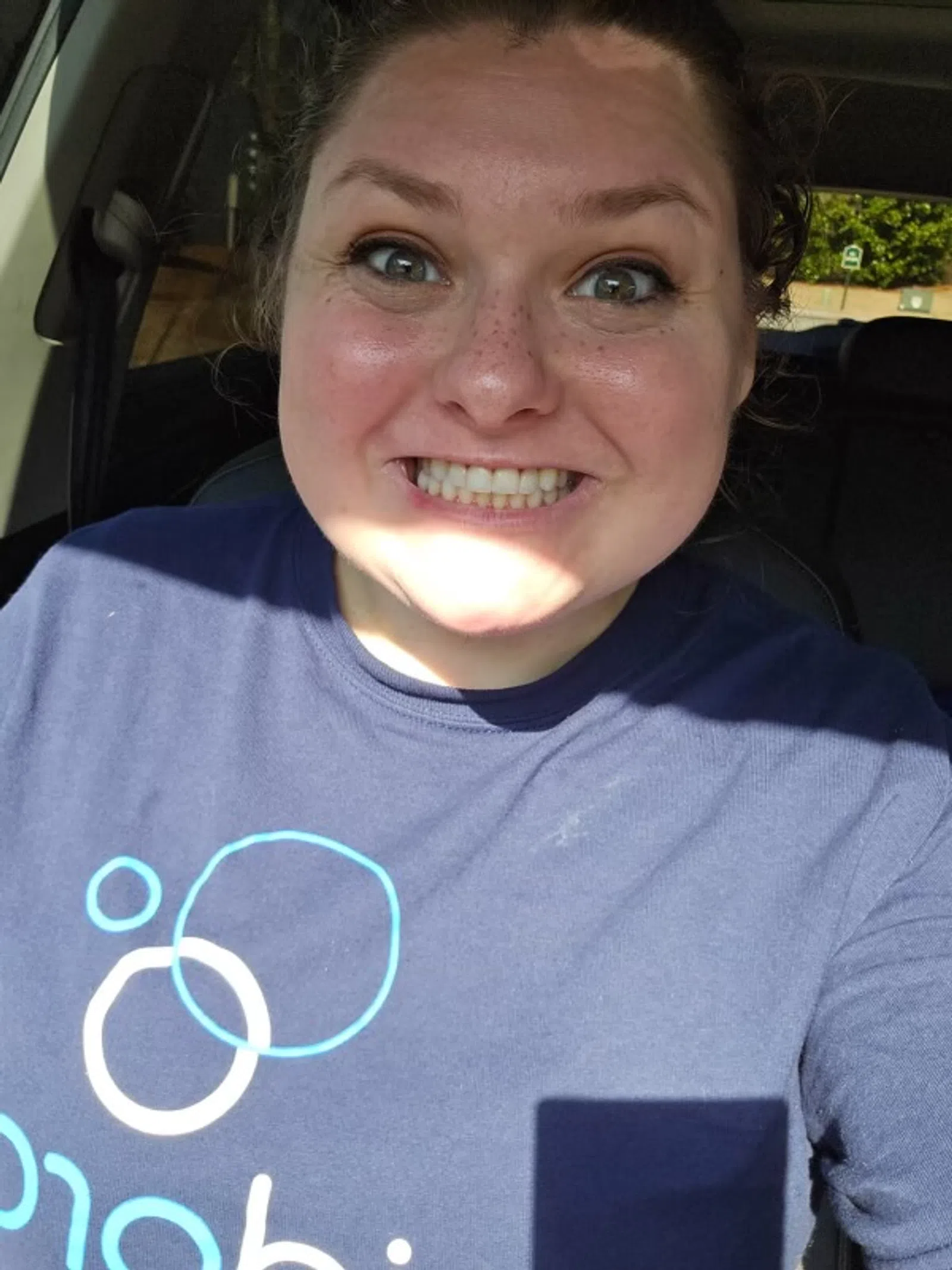 Smiling woman with freckles wearing a blue shirt with circular patterns sitting inside a car.