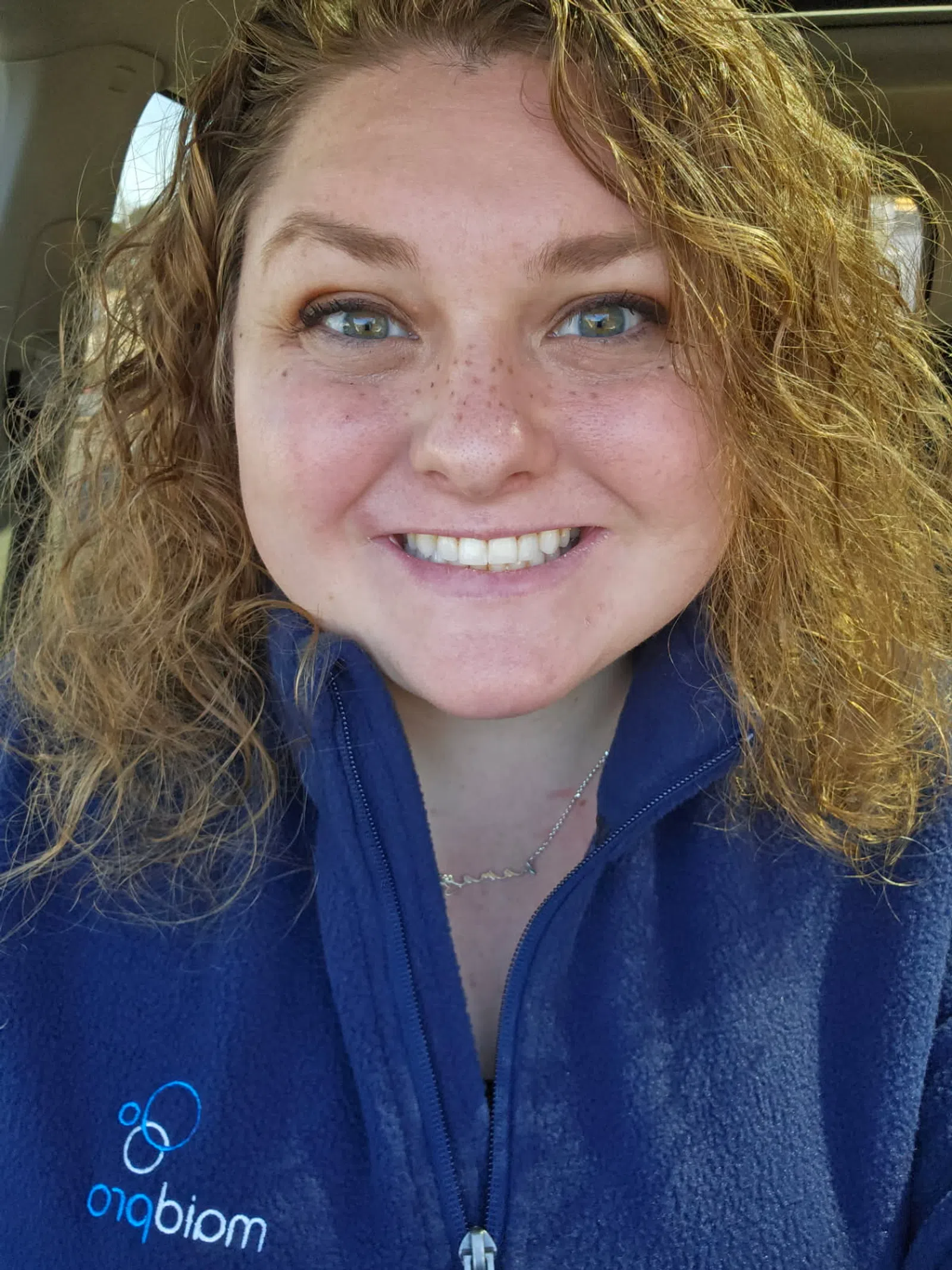 Smiling woman with curly hair wearing a blue zip-up jacket with a logo, sitting inside a car.