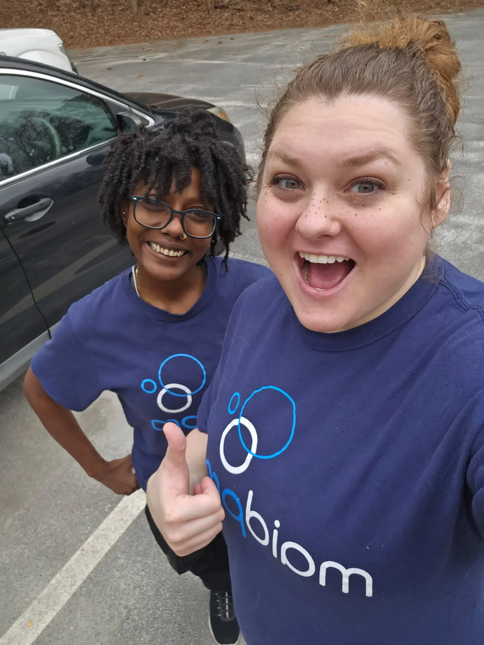 Two smiling women wearing matching blue Abiomed T-shirts, one giving a thumbs up, outdoors in a parking lot.