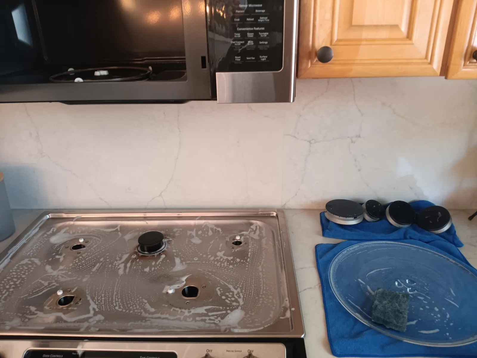 Clean stainless steel gas stove top and pot lids drying on blue towels in a kitchen with marble backsplash.