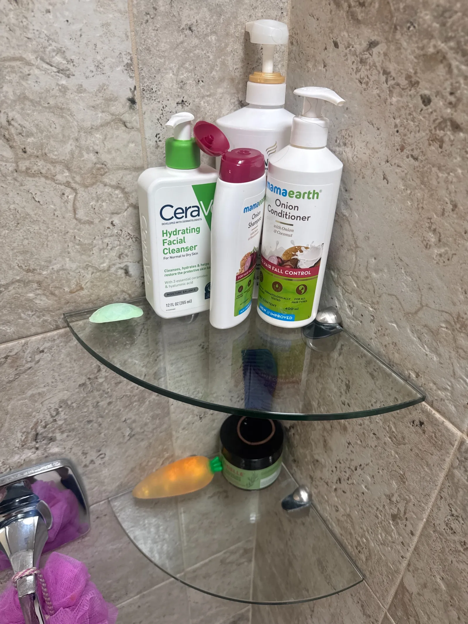 CeraVe cleanser and Mamaearth shampoo and conditioner bottles on glass shower corner shelves with tiled wall.