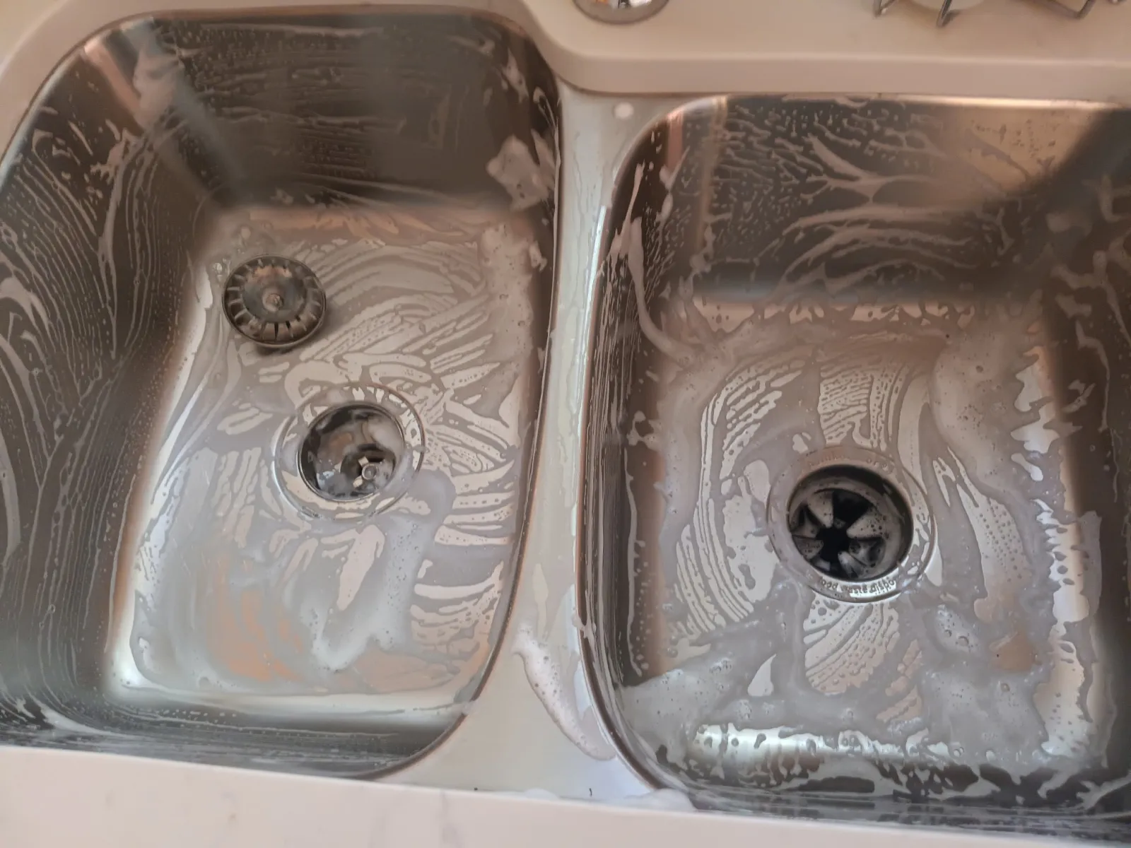 Two stainless steel kitchen sinks with soap suds and cleaning residue inside them.
