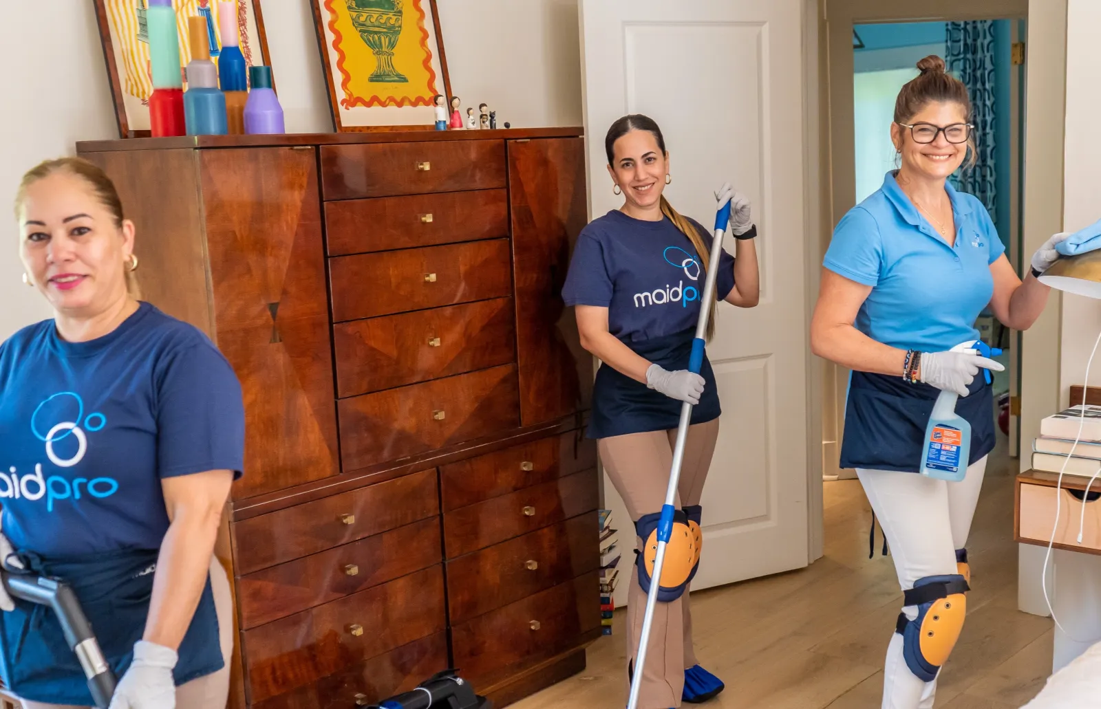 Three professional house cleaners wearing MaidPro uniforms cleaning a living room and smiling at the camera.