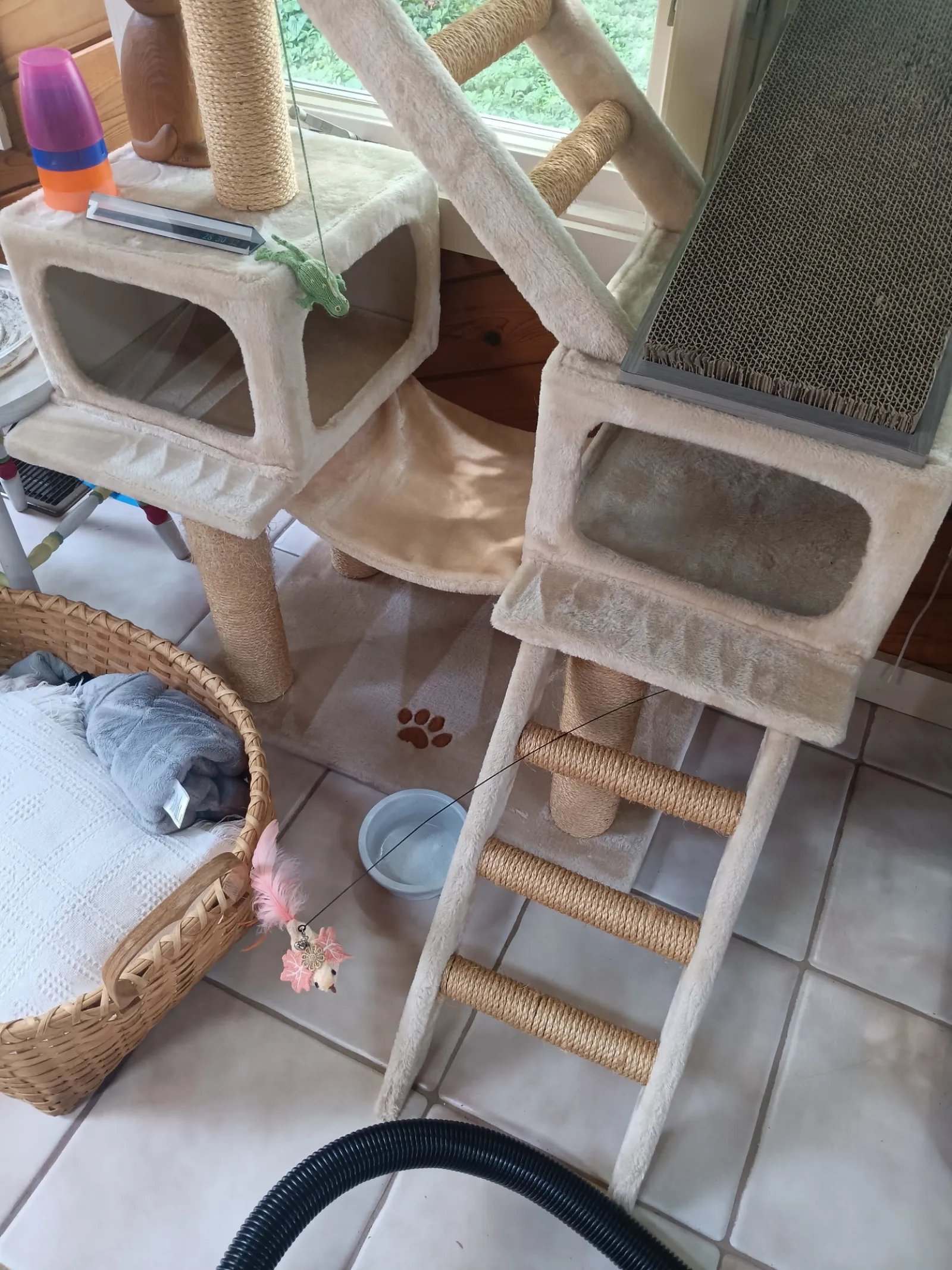 Beige multi-level cat tree with scratching posts, ramps, cozy boxes, and a cat toy with pink feathers indoors.