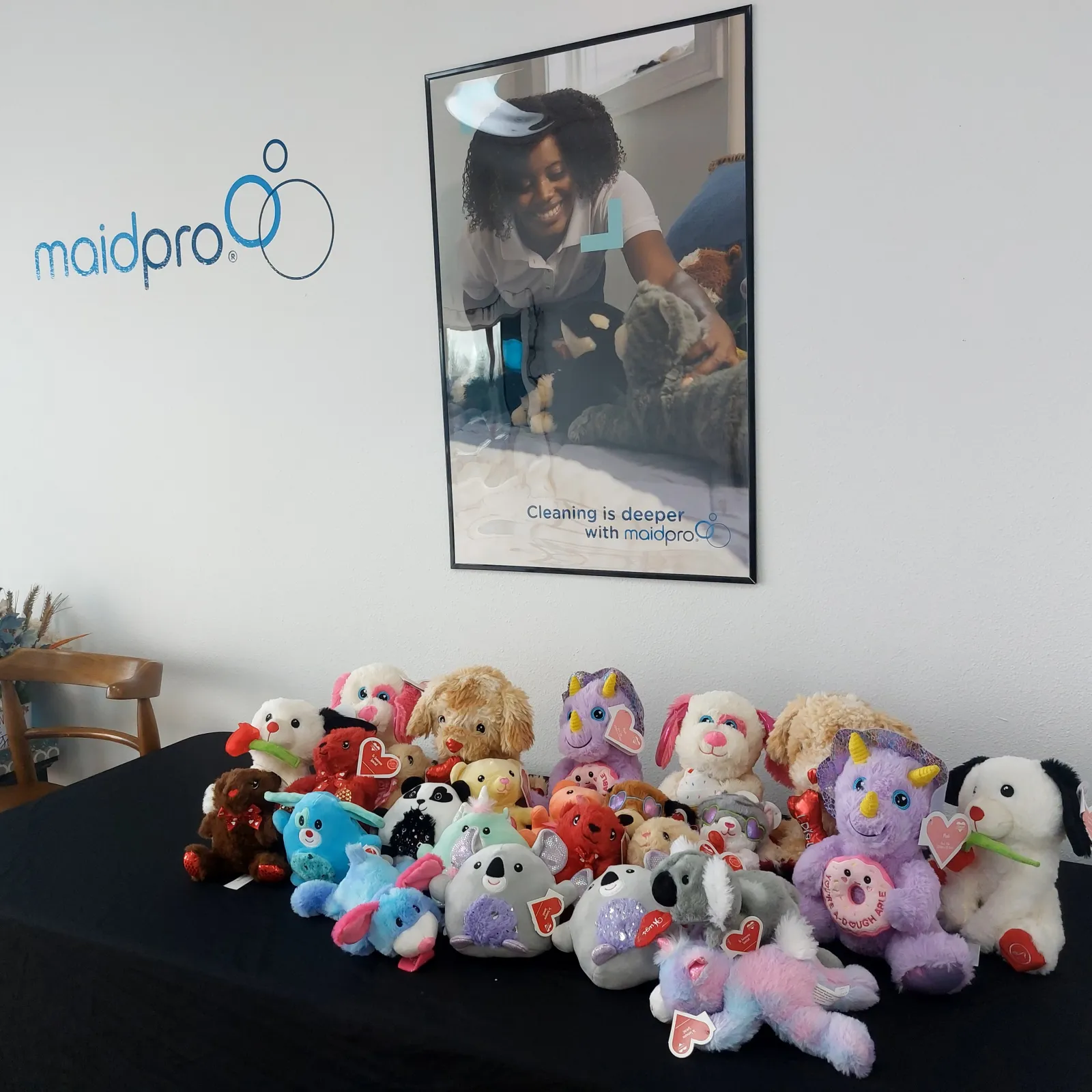 Variety of colorful stuffed animals displayed on a table under a MaidPro cleaning services poster and logo on the wall.