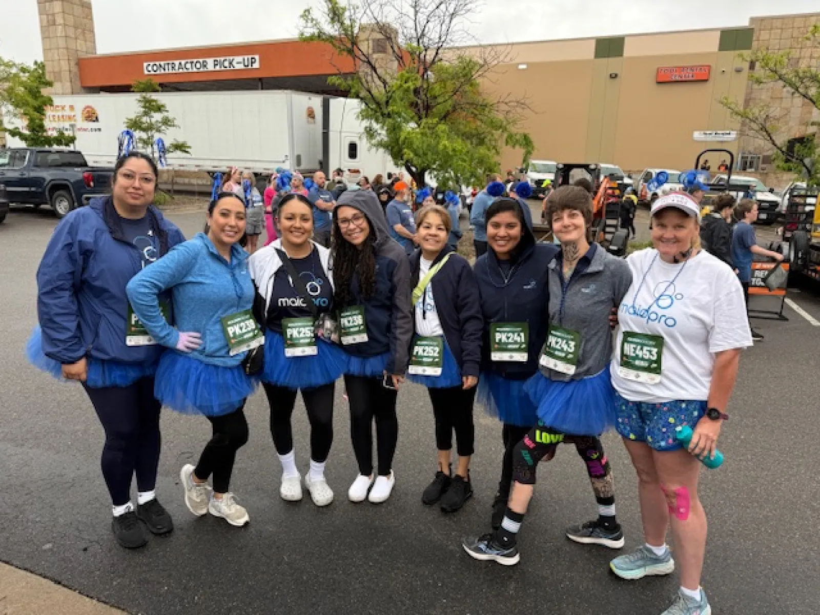 Group of women wearing blue tutus and race bibs standing together outdoors before a race event under cloudy sky.
