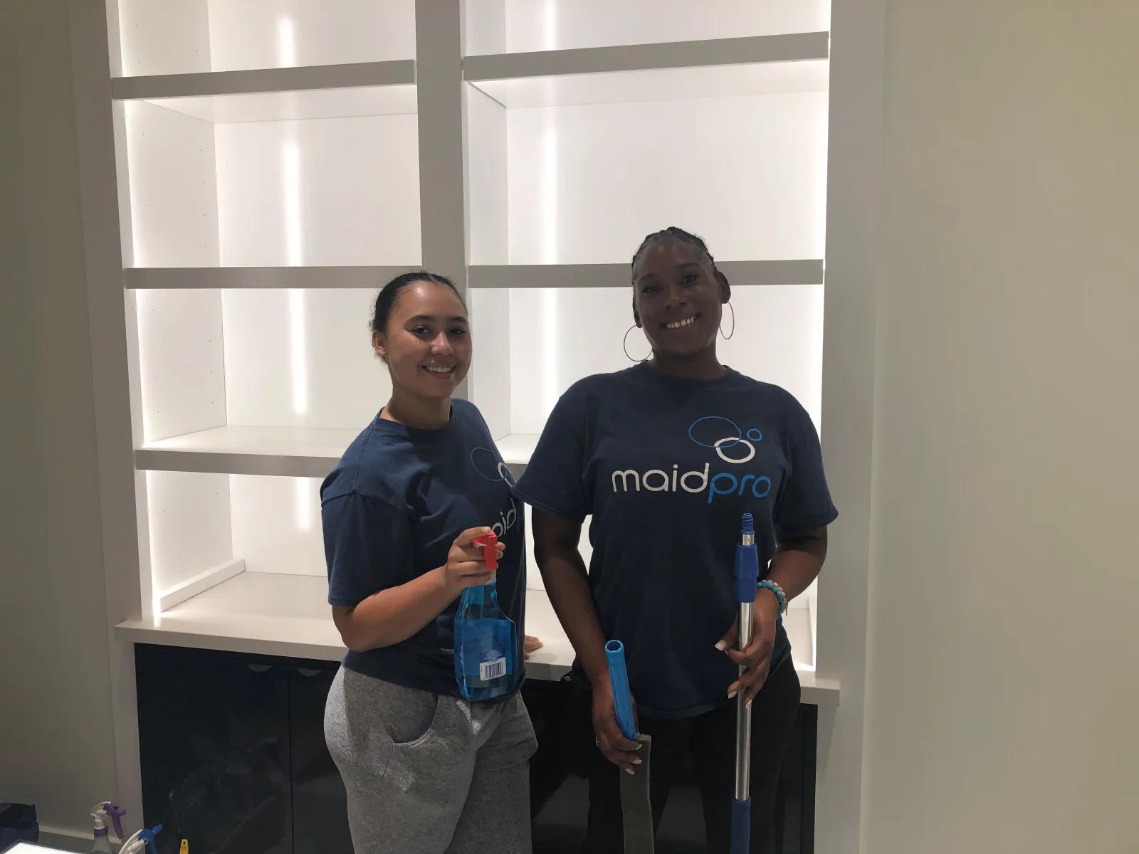 Two women in Maid Pro shirts holding cleaning supplies in a modern, empty white shelving area.