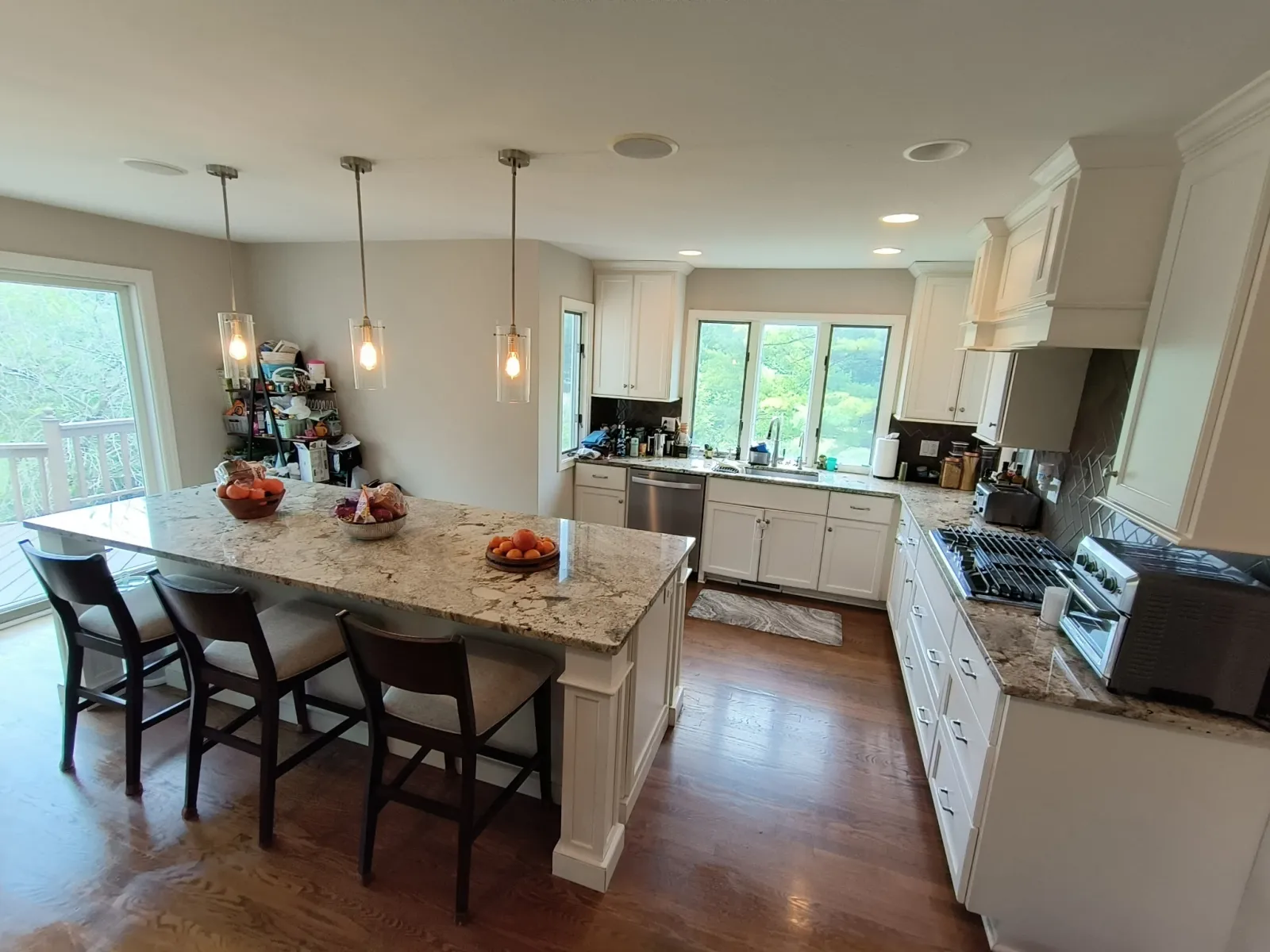 Bright modern kitchen with white cabinets, granite island, hardwood floors, and large windows overlooking greenery.
