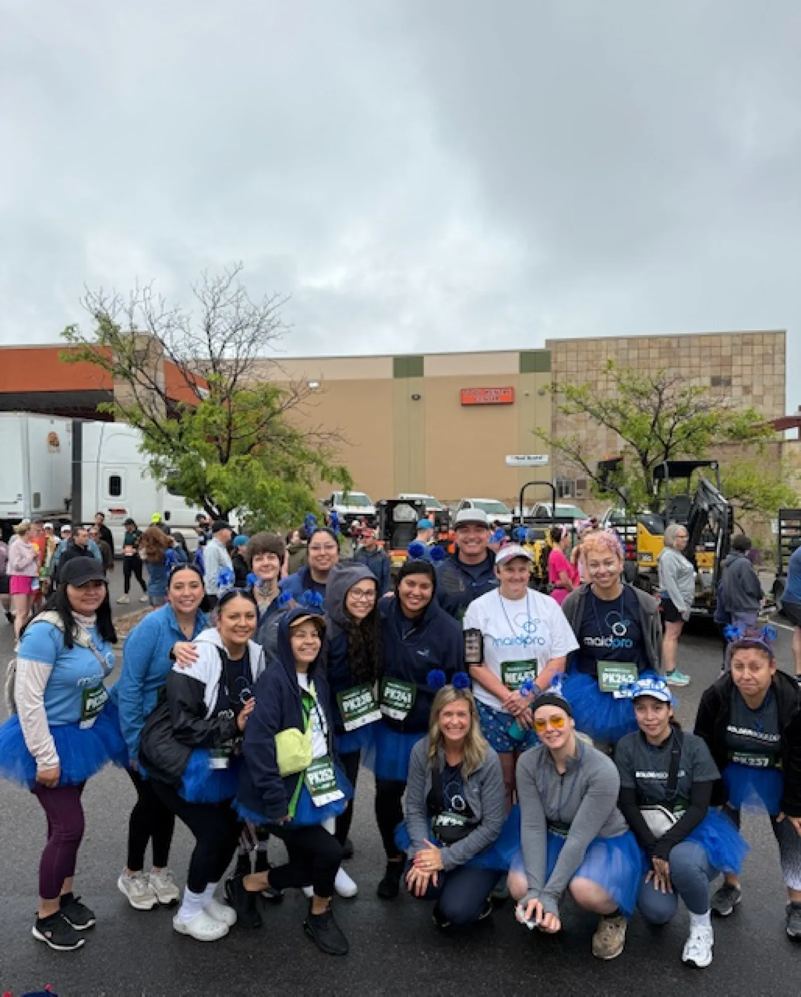 Group of smiling participants in blue tutus and race bibs posing outdoors before a community race event on a cloudy day.