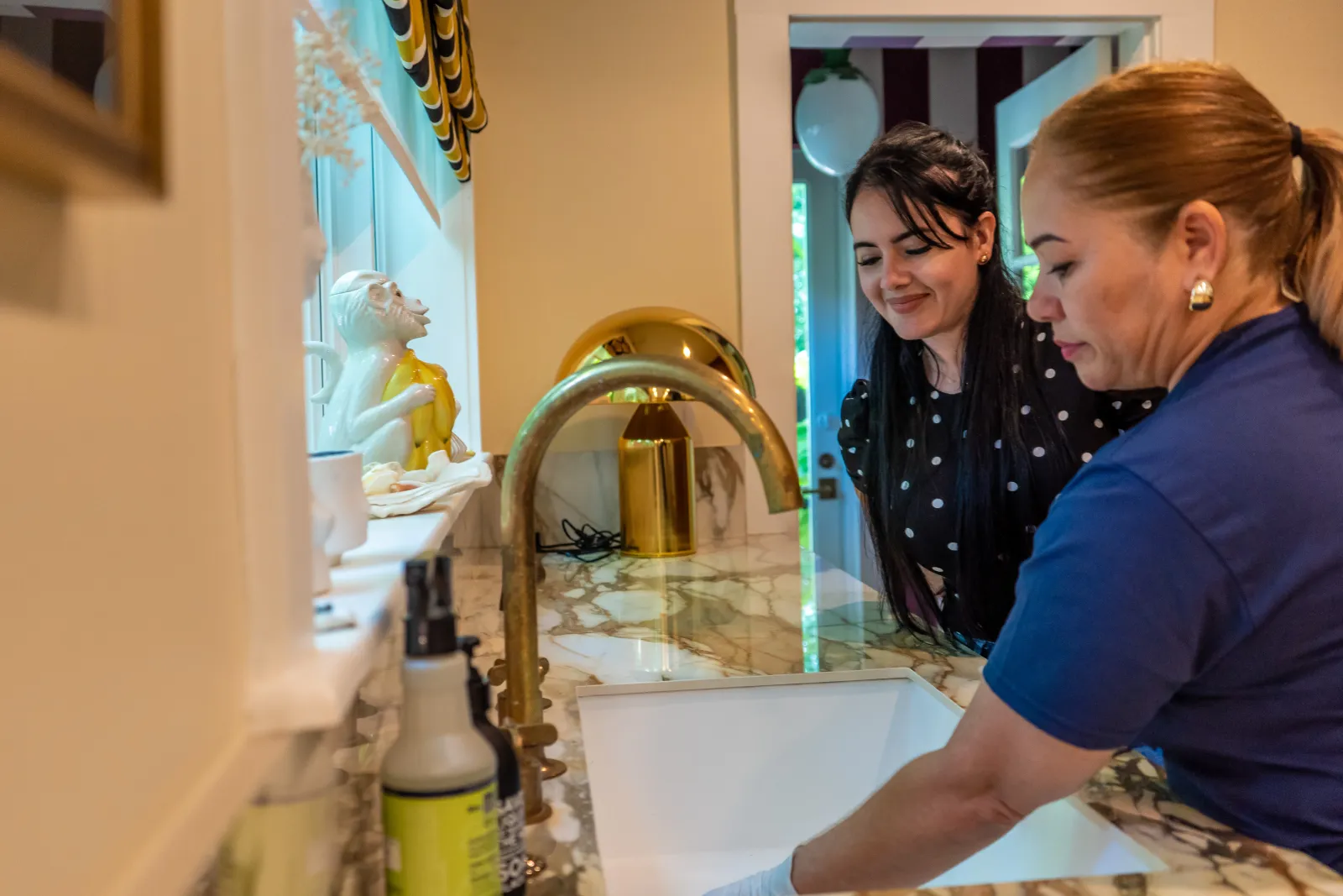 Two women smiling while cleaning a modern kitchen sink with gold faucet and marble countertop