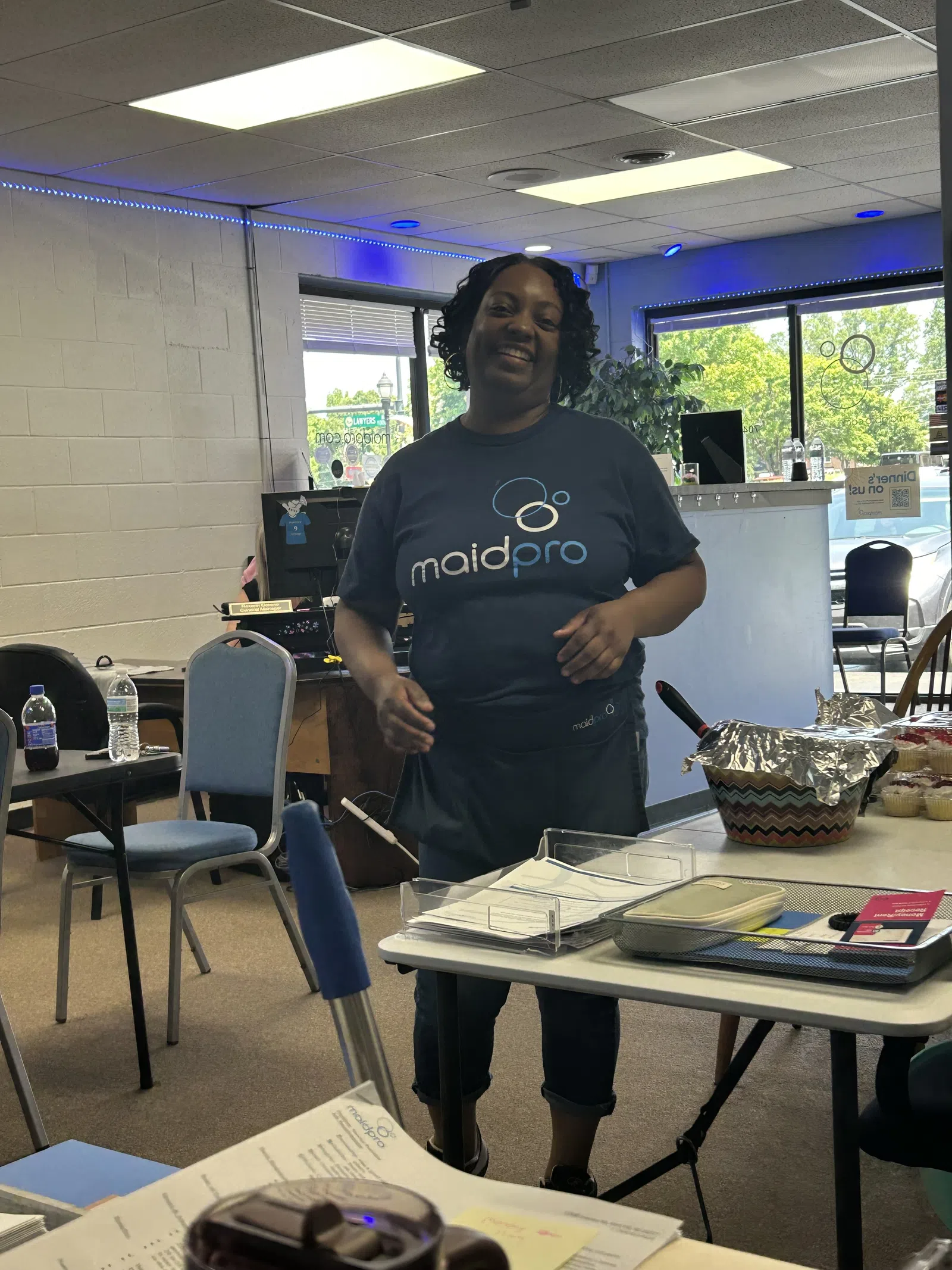 Smiling woman wearing a MaidPro t-shirt standing in a room with tables, chairs, and office supplies.