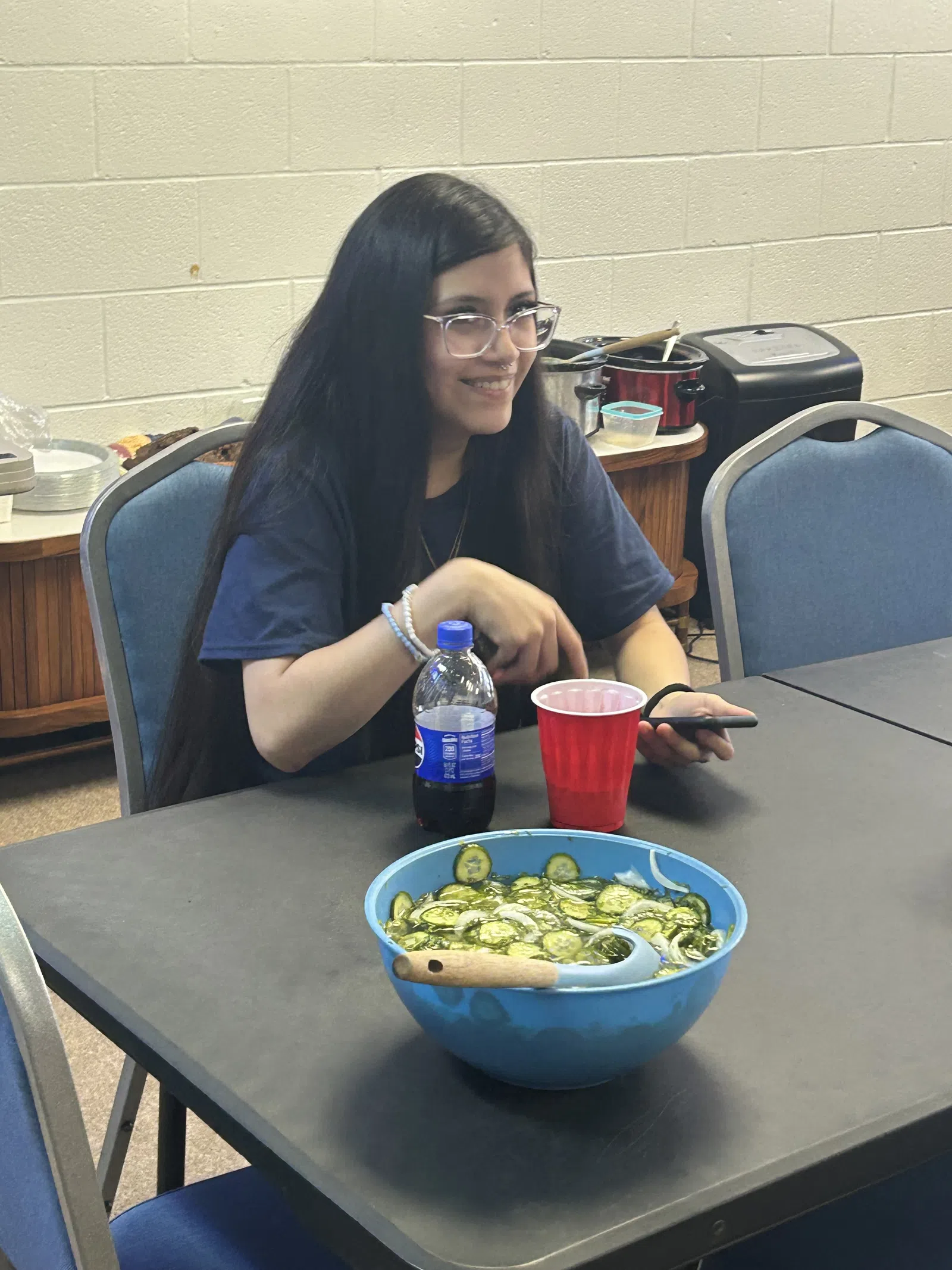 Young woman with long dark hair sitting at a table with a bowl of sliced cucumbers, red cup, and soda bottle.