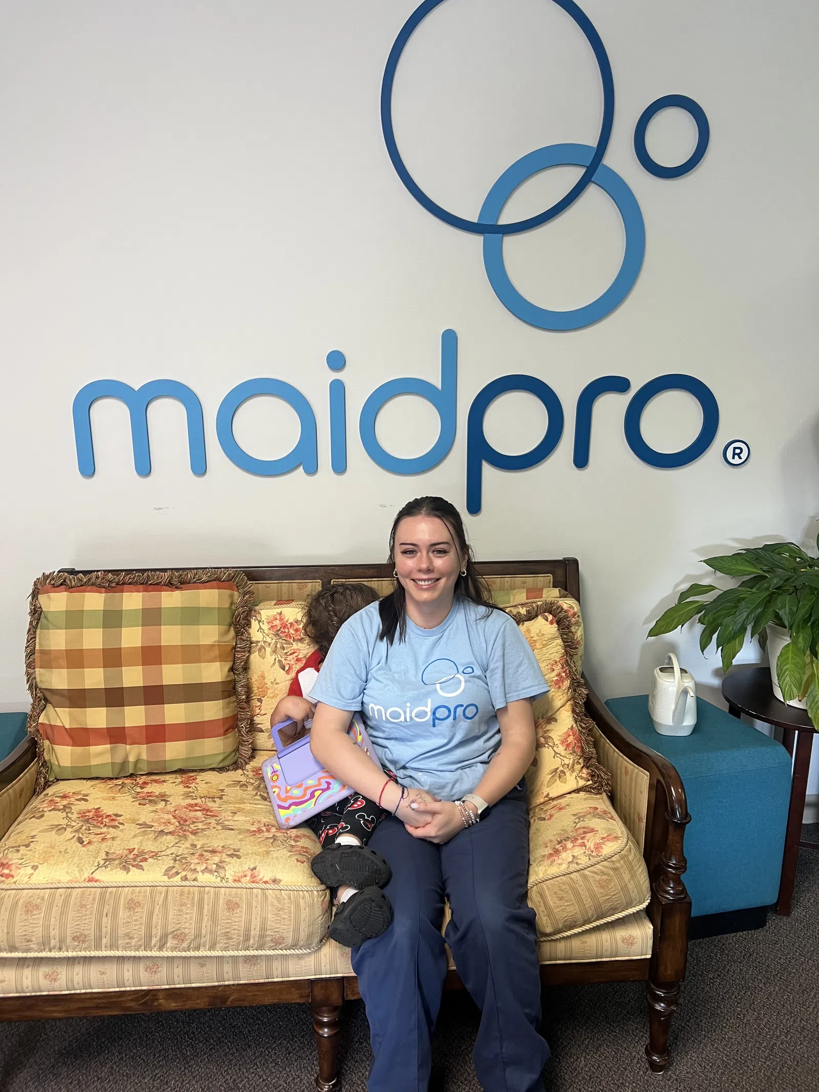 Smiling woman in maidpro shirt sitting on vintage sofa with child holding a toy, maidpro logo on wall behind.