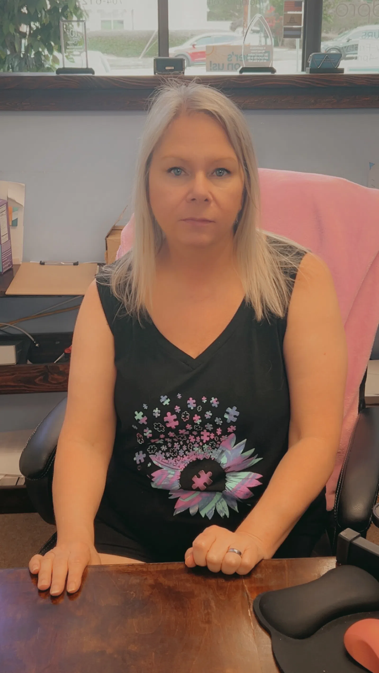 Woman with blonde hair wearing a black sleeveless shirt with colorful puzzle flower design sitting at a wooden desk.