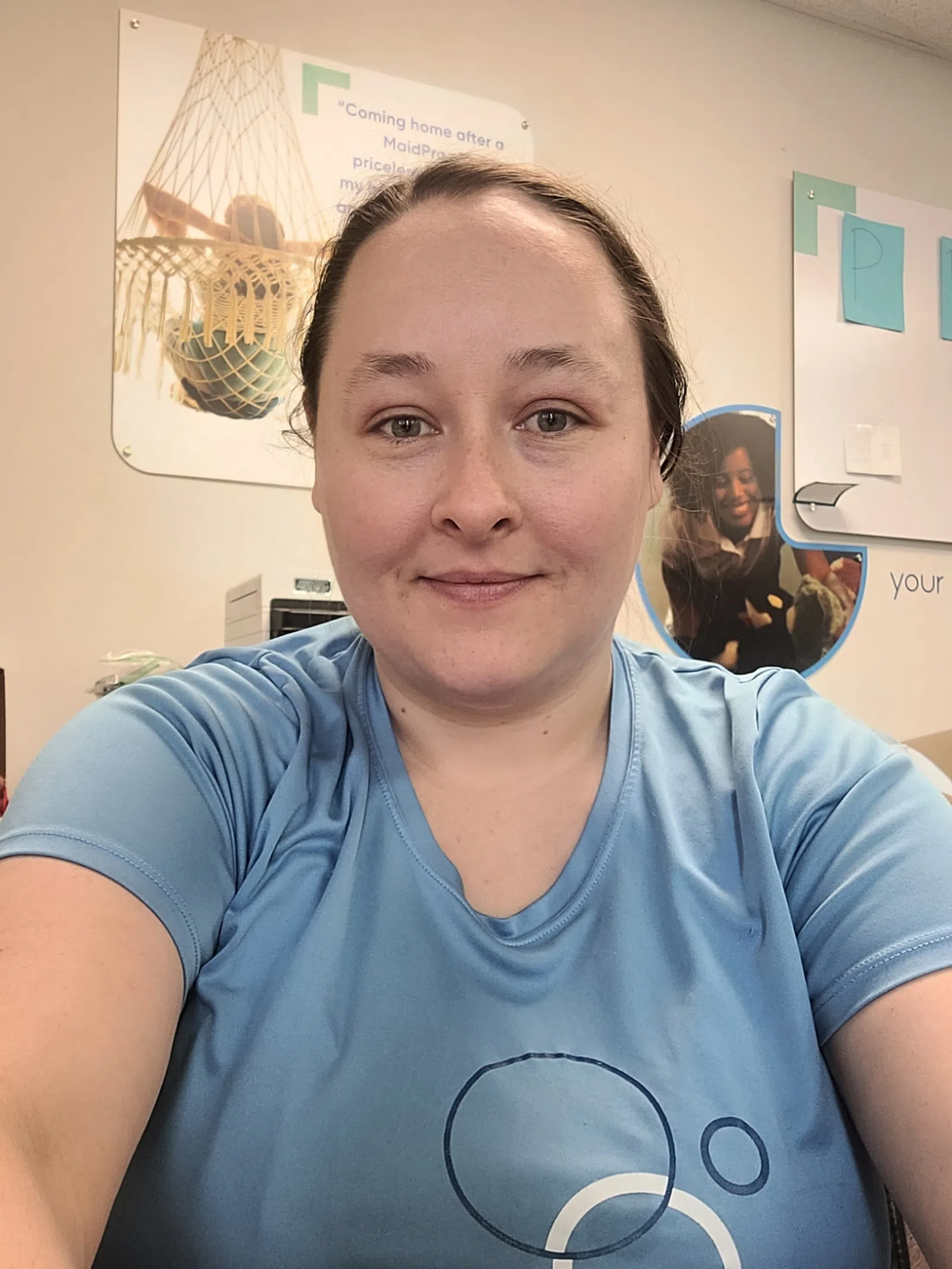 Smiling woman in a blue shirt taking a selfie in an office with posters and bulletin boards in the background