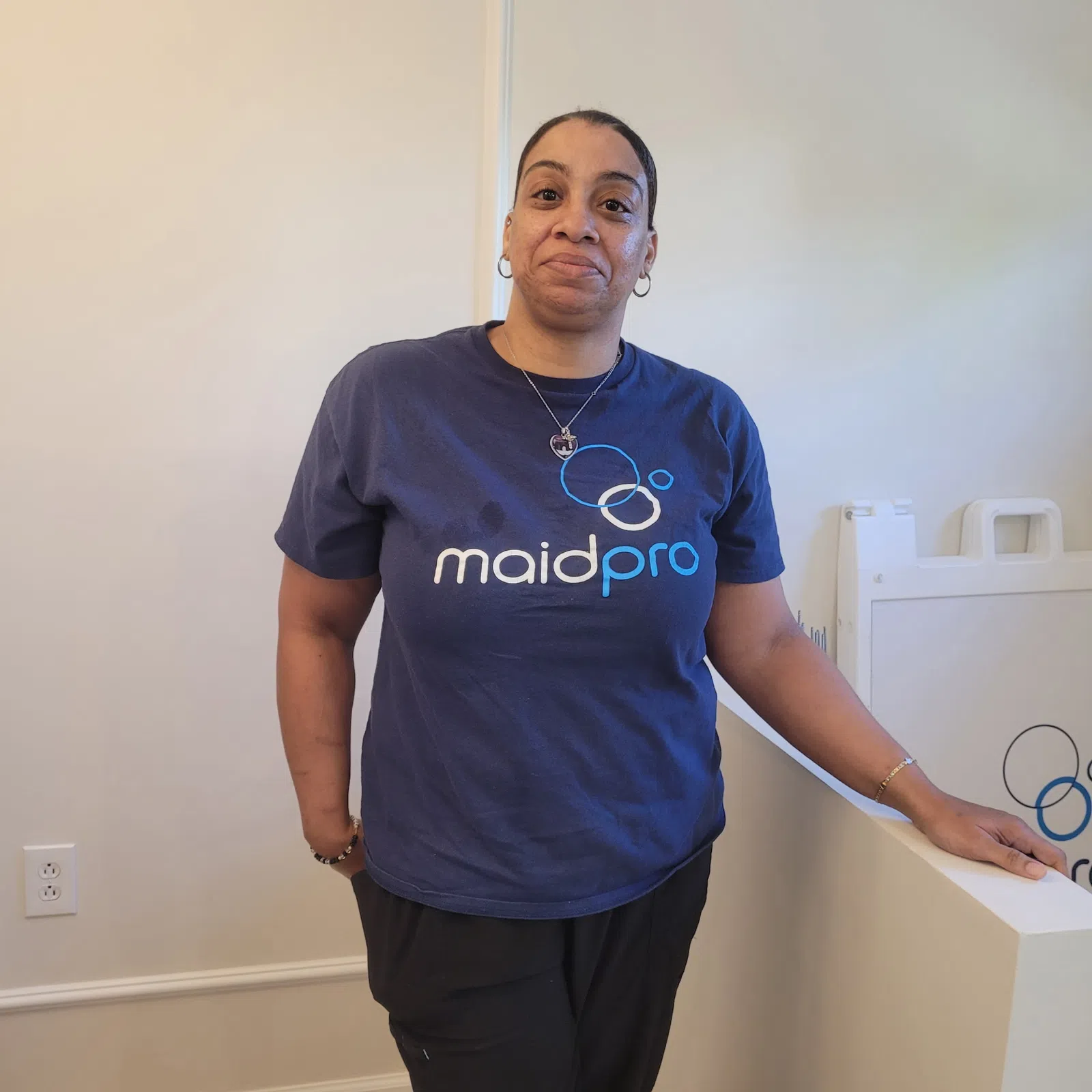 Woman in a navy MaidPro shirt standing indoors beside a white partition with cleaning supplies visible.