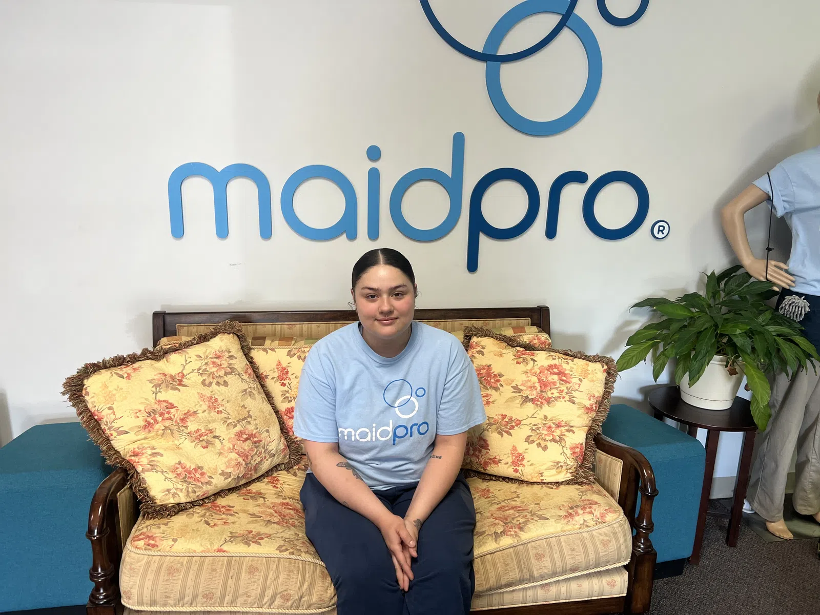Maidpro employee sitting on floral couch in front of Maidpro logo wearing blue uniform shirt.