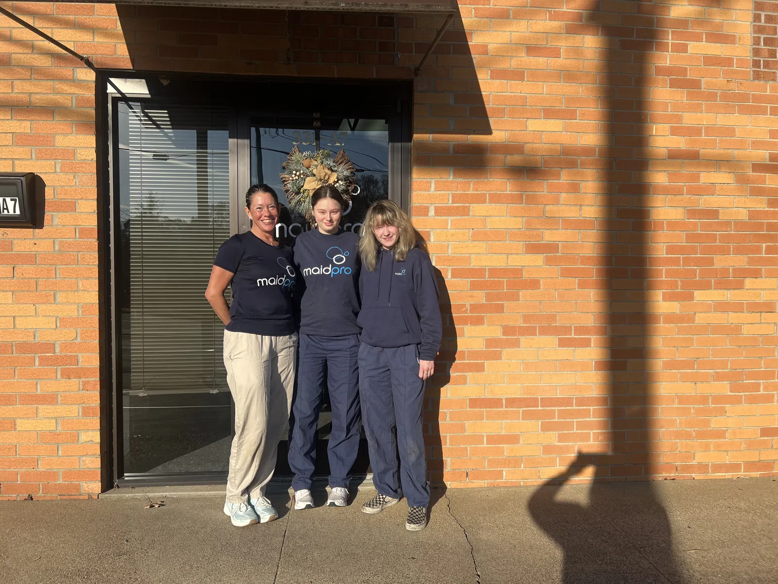 Three women in MaidPro uniforms standing outside a brick building with a glass door and wreath.