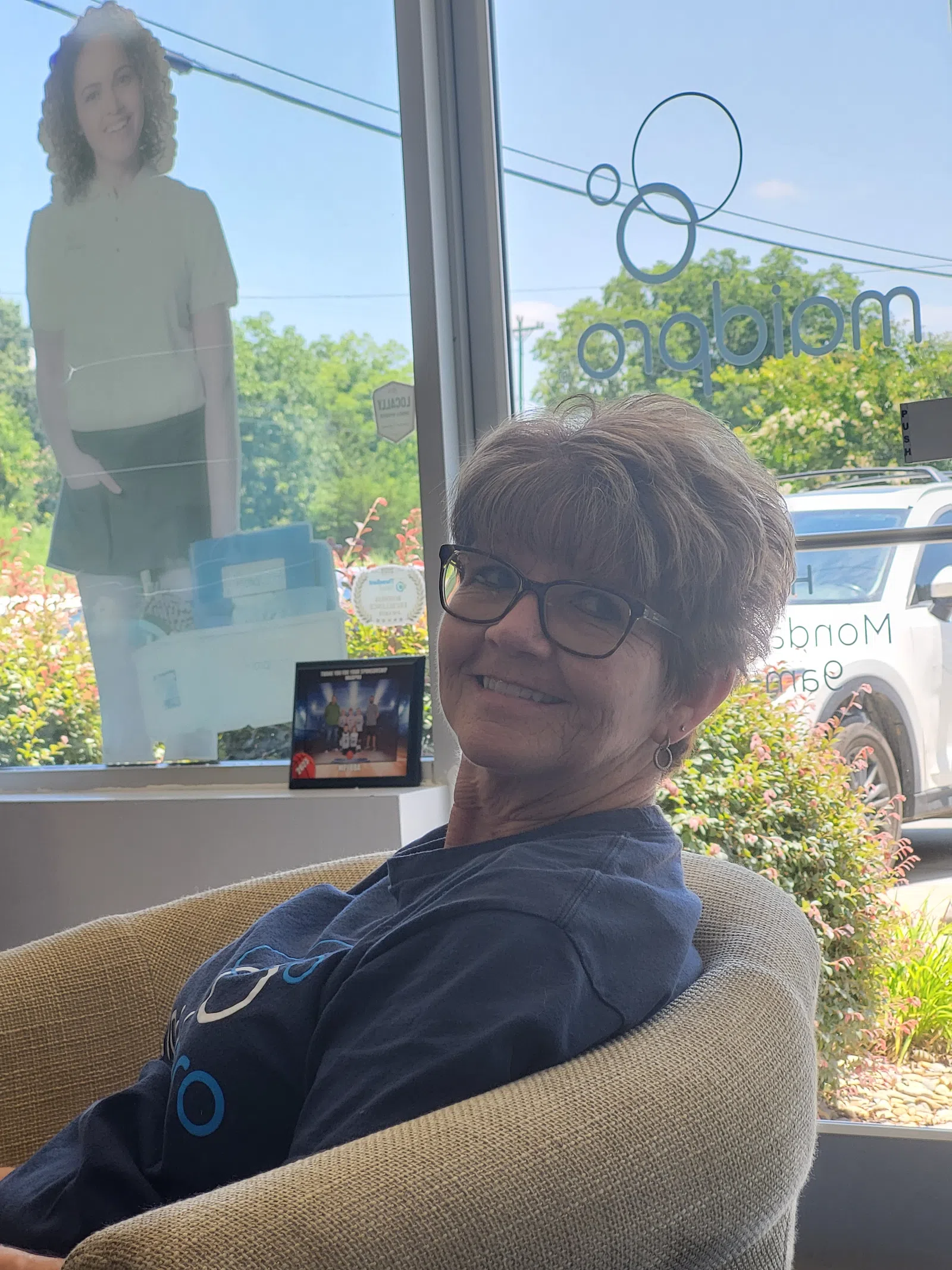 Smiling woman wearing glasses sitting in a chair near a window with outdoor greenery and a standing cutout figure visible.