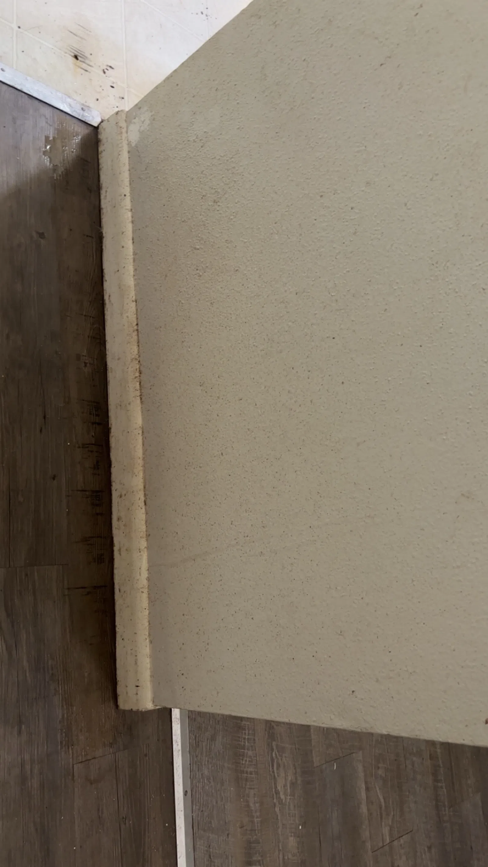 Corner of a beige wall meeting a wooden floor with a white baseboard and tile flooring above it.