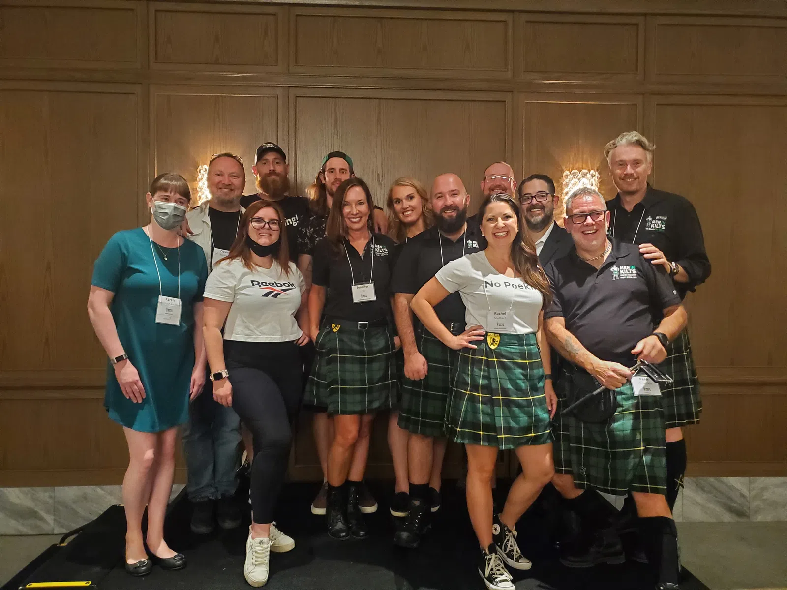 Group of eleven people smiling indoors, most wearing green tartan kilts and casual tops, posing in front of wood panel wall.
