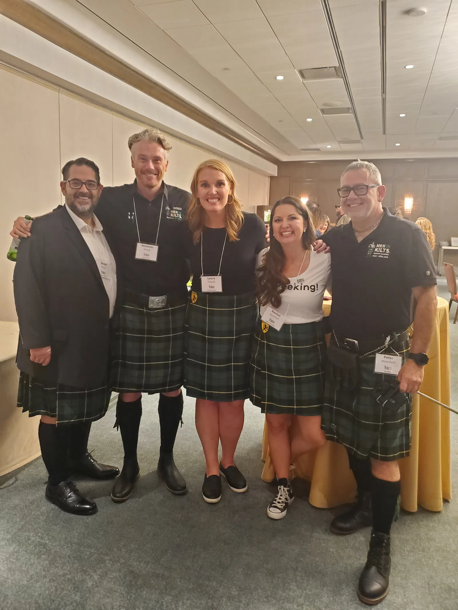 Five people smiling indoors wearing matching green tartan kilts and name badges at an event.