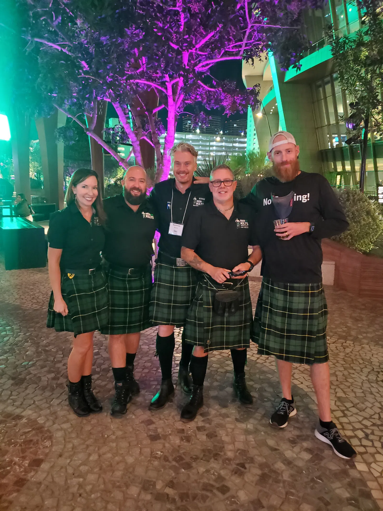 Group of five people wearing green plaid kilts and black tops posing outdoors at night with colorful tree lighting.