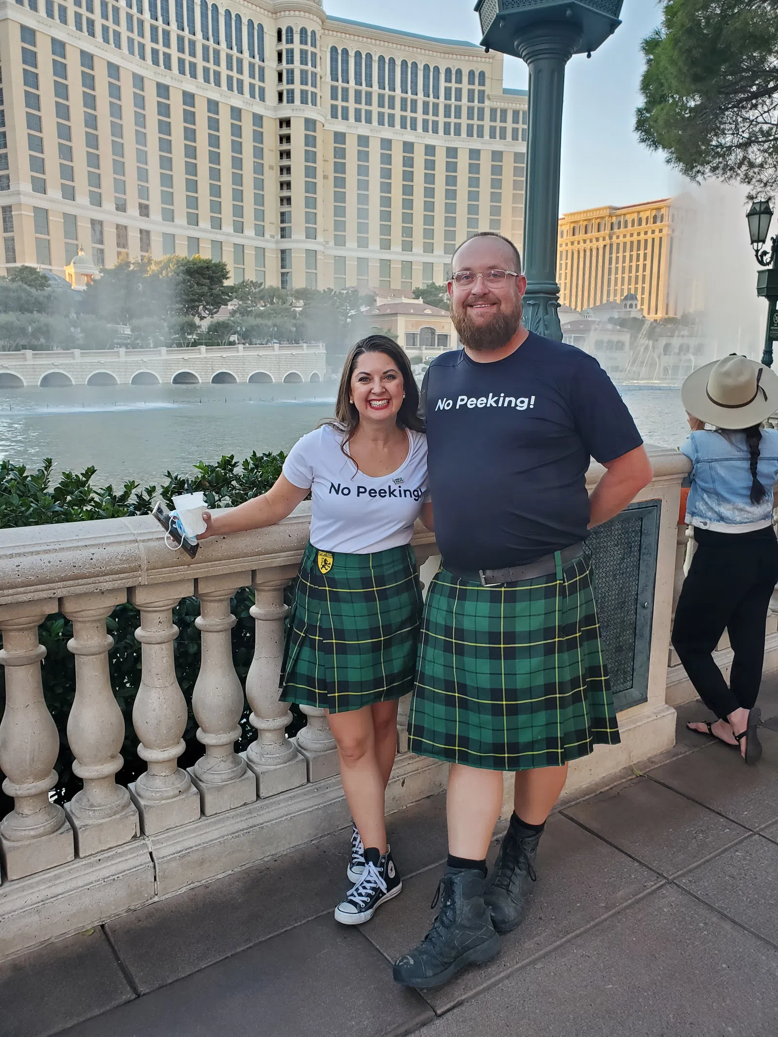Smiling couple wearing matching green plaid kilts and No Peeking shirts by a fountain with hotel buildings in the background