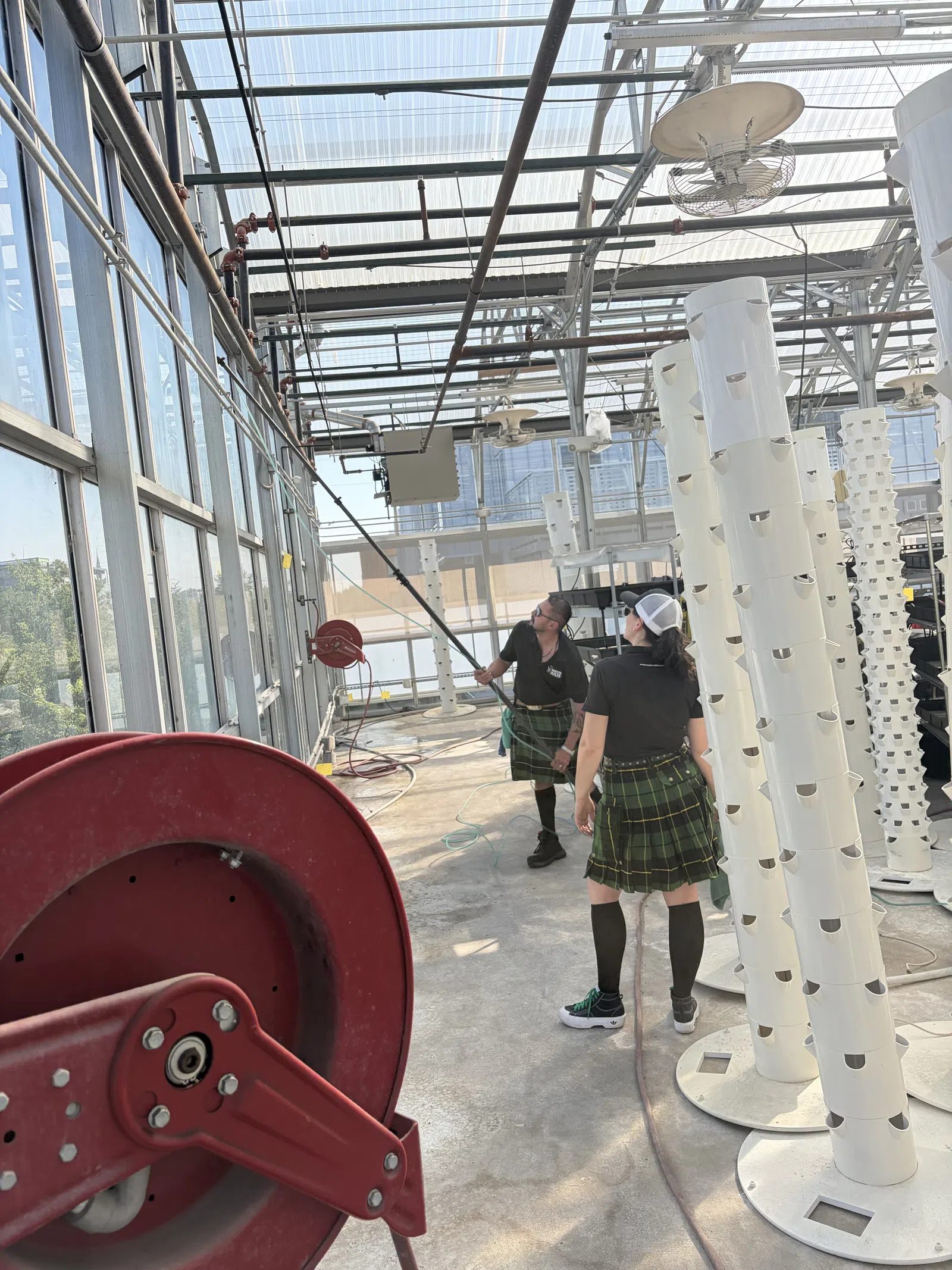 Two people in kilts inside a greenhouse with vertical hydroponic towers and red hose reel.