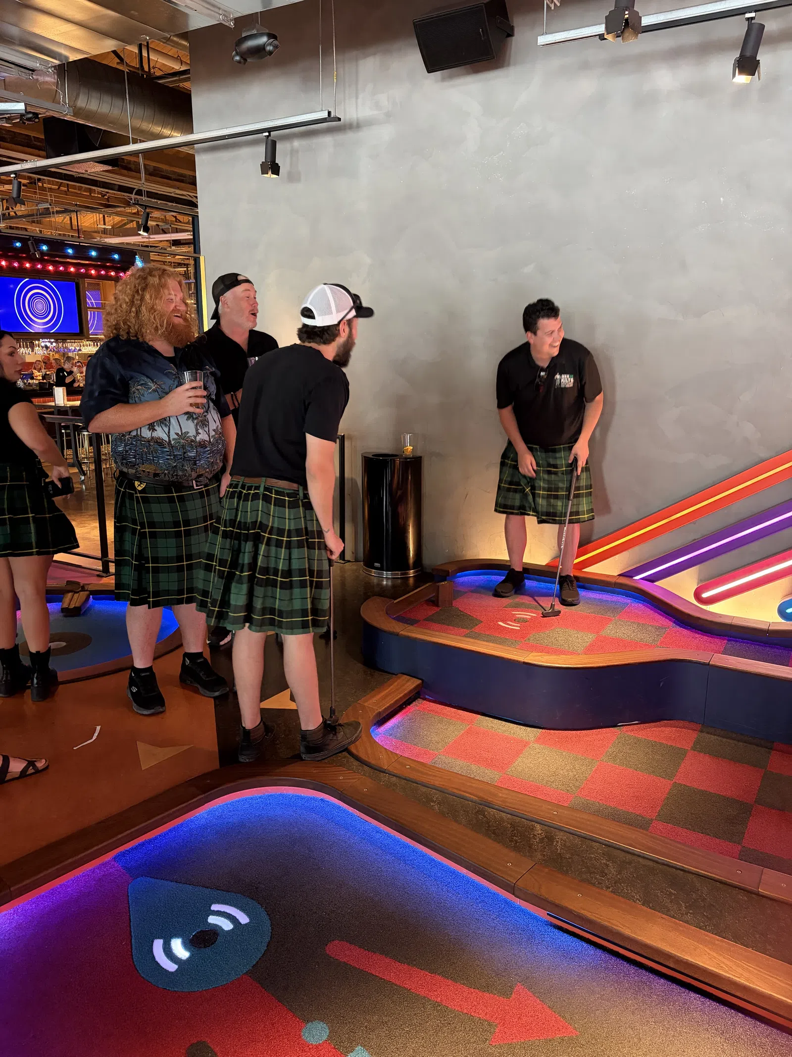 Group of adults wearing kilts playing indoor mini golf in a colorful, modern entertainment venue.
