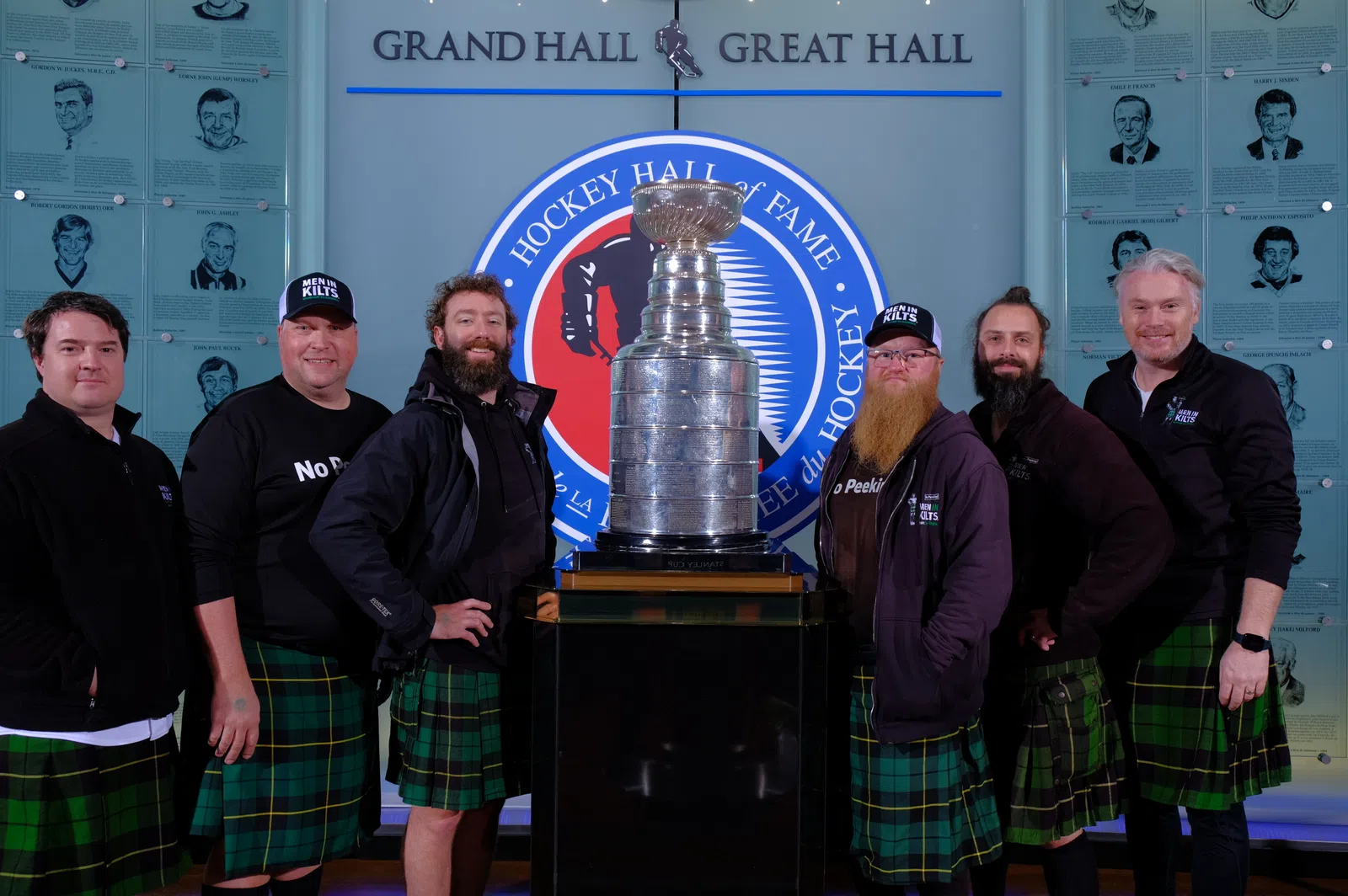 Group of men in green plaid kilts posing around the Stanley Cup in the Hockey Hall of Fame Grand Hall.