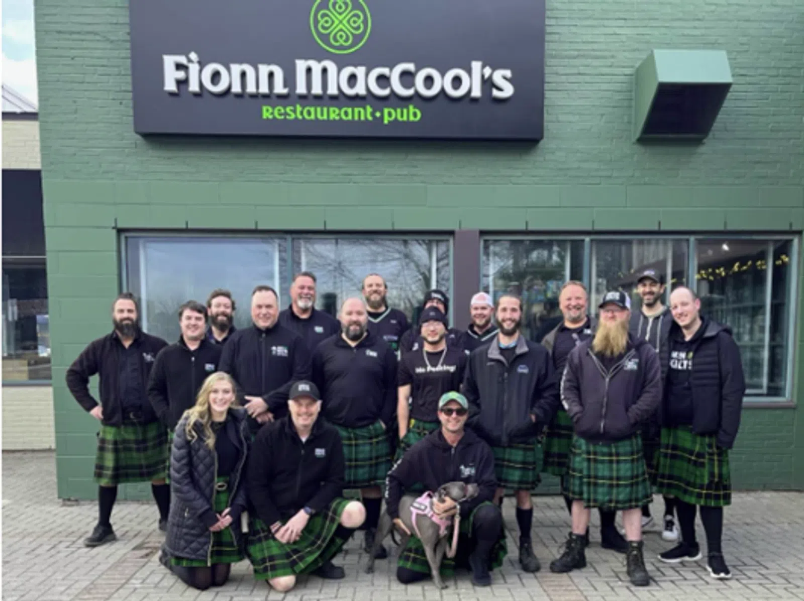 Group of people wearing green plaid kilts posing outside Fionn MacCool's restaurant and pub on a city sidewalk.